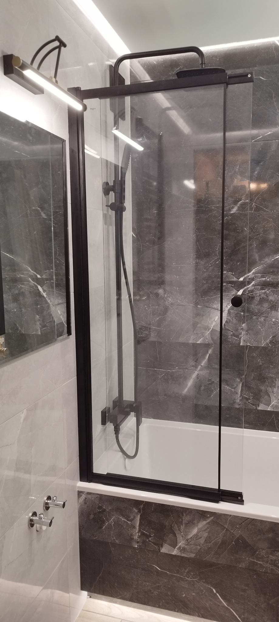 Kabina prysznicowa z czarnymi profilami i armaturą, zamontowana nad wanną, oświetlona lampą nad lustrem, w łazience z szarymi płytkami imitującymi marmur.