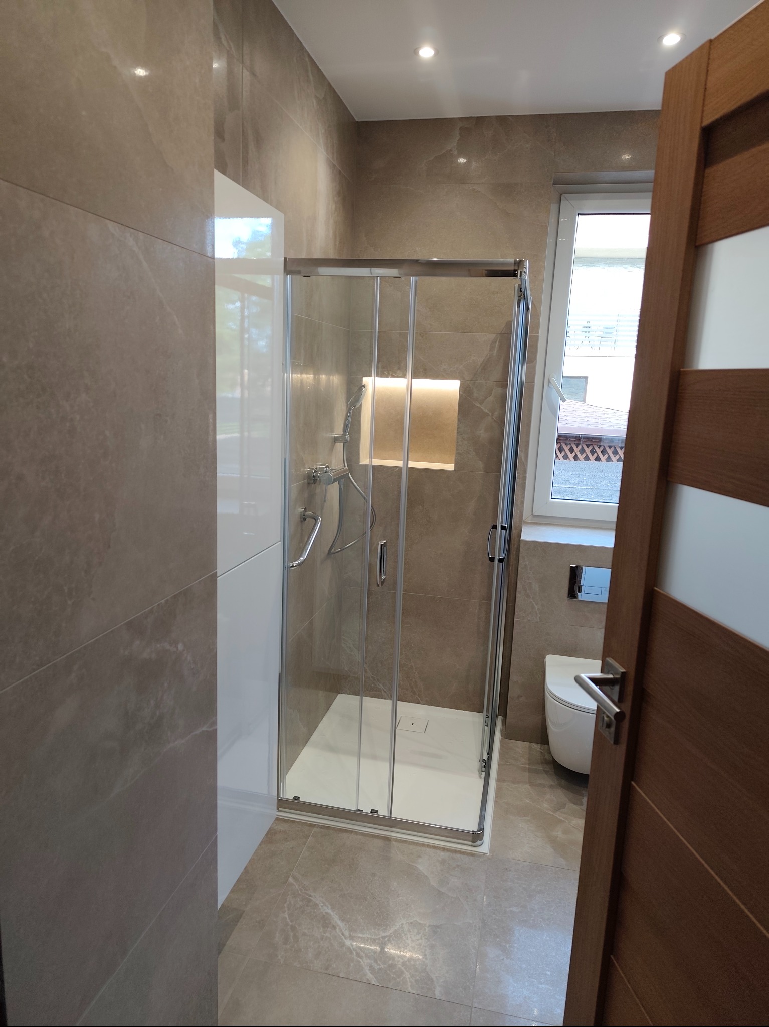 Beżowa łazienka z kabiną prysznicową walk-in. Widoczne płytki imitujące marmur, podświetlona wnęka i częściowo otwarta drewniana rama drzwi.