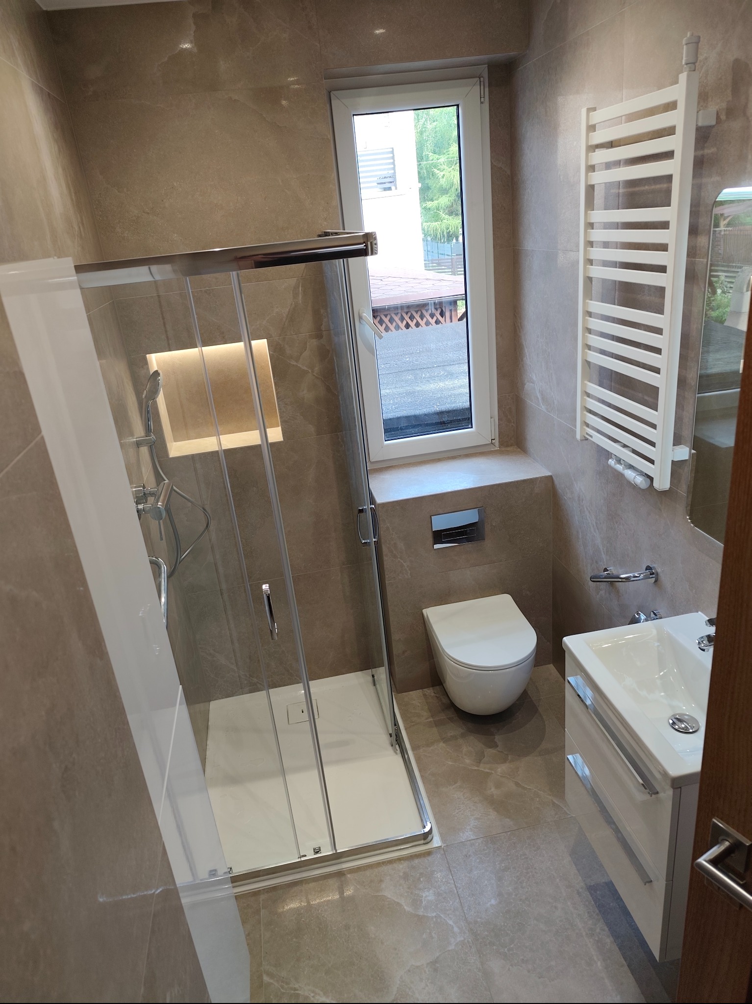 Jasna łazienka z kabiną prysznicową typu walk-in, beżowe płytki imitujące marmur, biała umywalka z szafką i grzejnik drabinkowy. Okno z widokiem na zewnątrz.