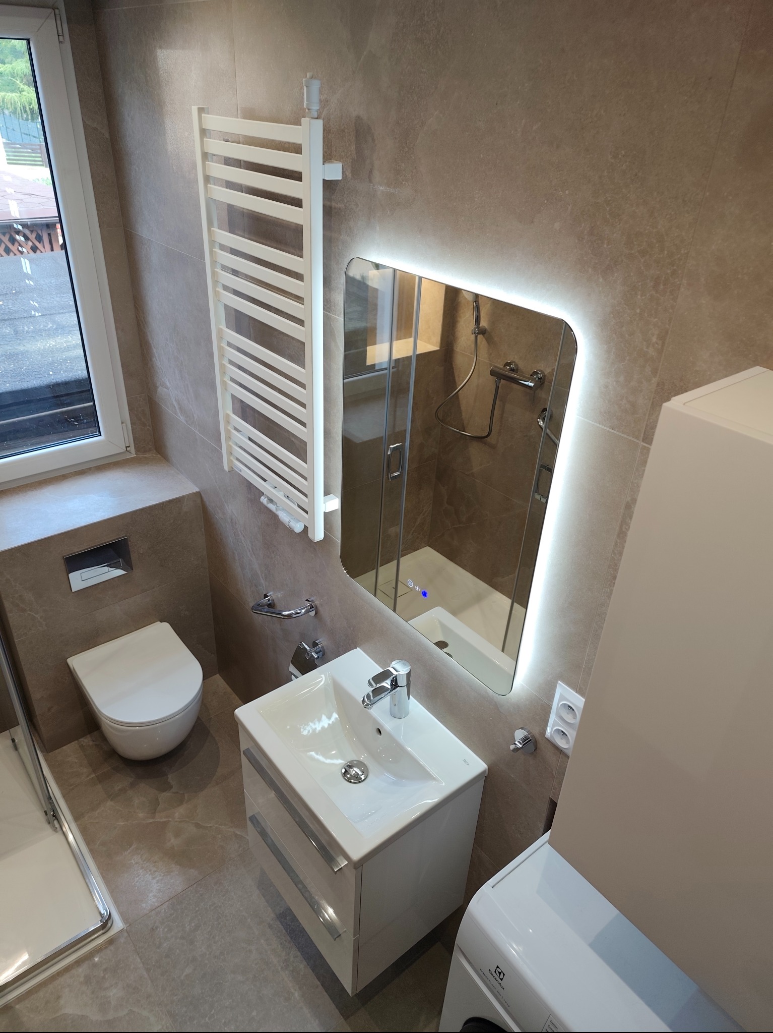 Nowoczesna łazienka w beżowej tonacji: podświetlane lustro, biała umywalka z szafką, grzejnik drabinkowy i kabina prysznicowa z widocznym odpływem liniowym.