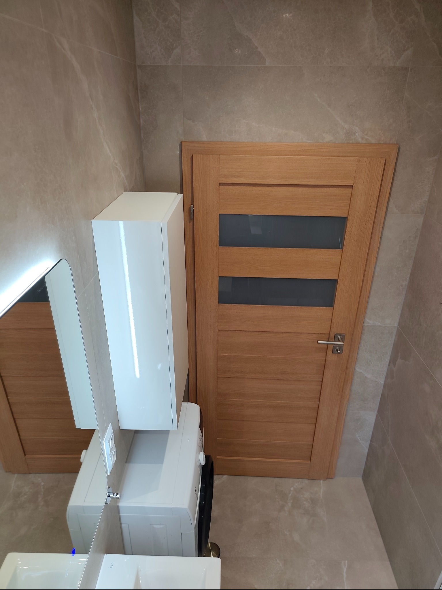 Nowoczesna łazienka z beżowymi płytkami imitującymi marmur, drewnianymi drzwiami z przeszkleniami i białą szafką wiszącą nad pralką. Lustro z podświetleniem.