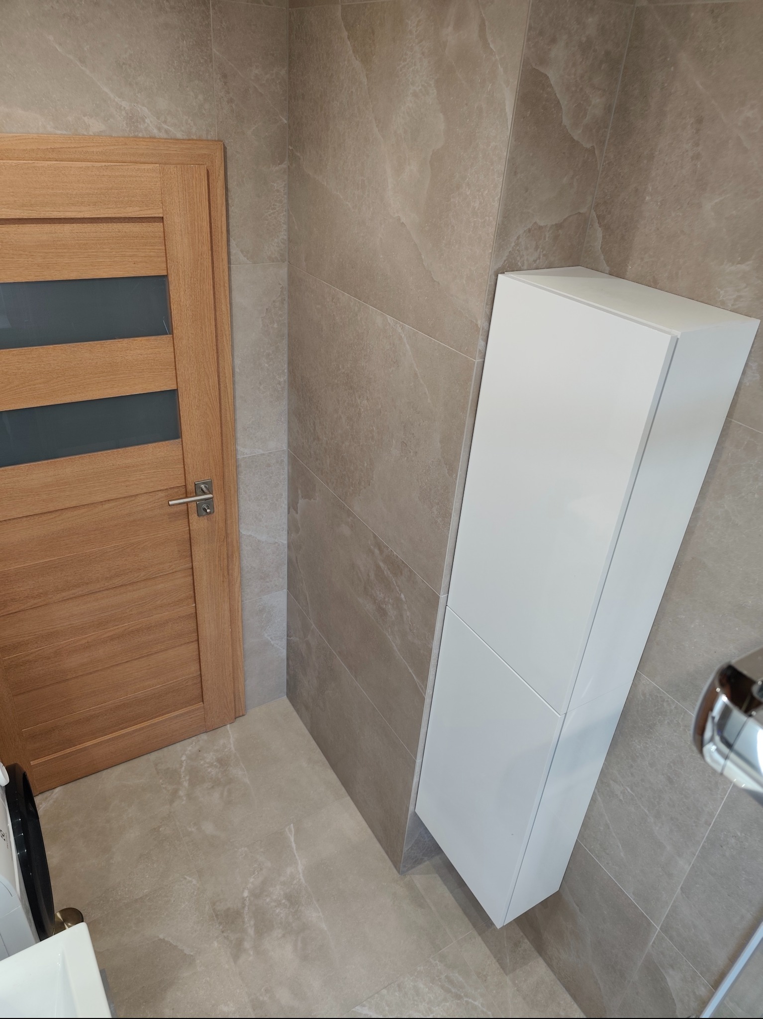 Wykończona łazienka z beżowymi płytkami imitującymi marmur, drewnianymi drzwiami z przeszkleniem i białą, wąską szafką. Nowoczesny design.