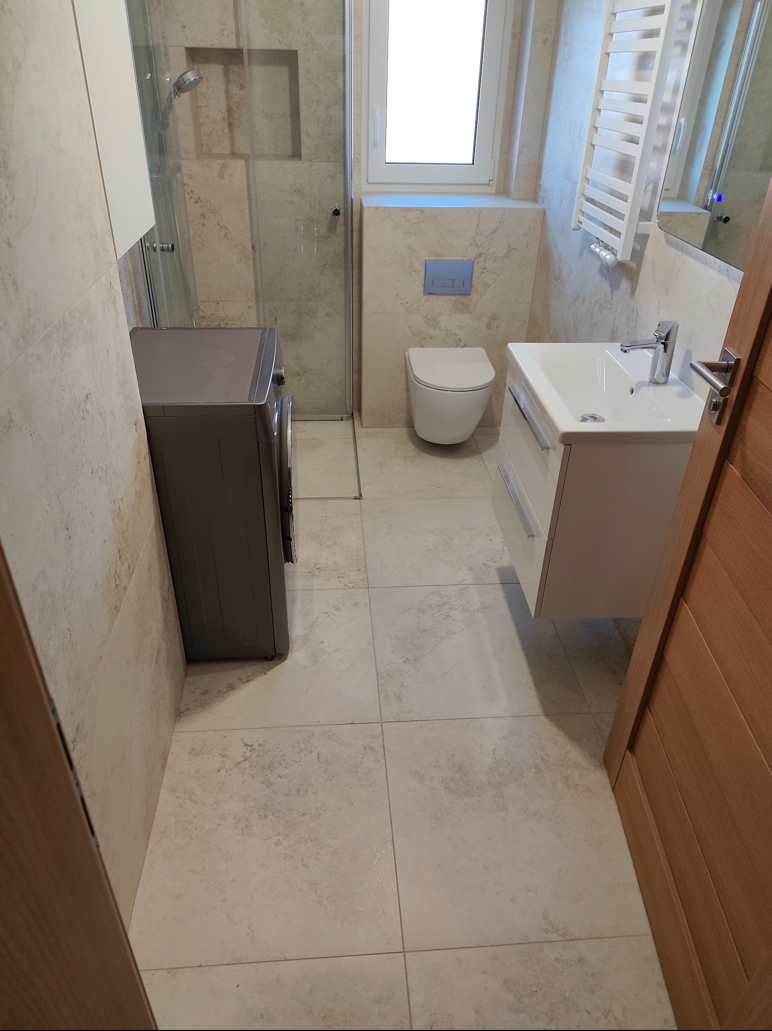 Wykończona łazienka z beżowymi płytkami, kabiną prysznicową, pralką, umywalką z szafką i grzejnikiem drabinkowym. Minimalistyczny styl, jasne wnętrze.