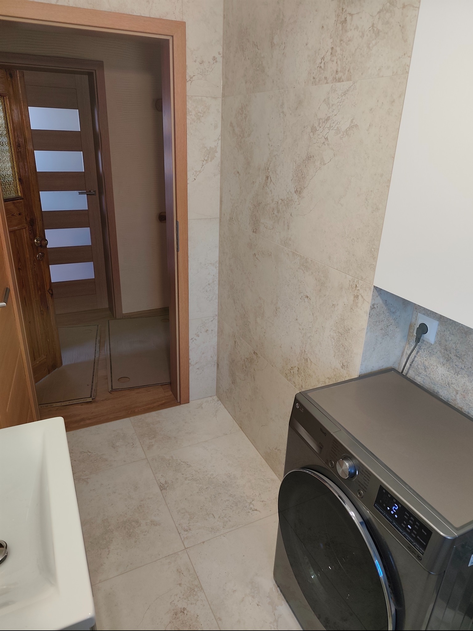 Jasne, marmurkowe płytki w wykończonej łazience z pralką LG i widokiem na otwarte drzwi z drewnianą ościeżnicą. Neutralna kolorystyka wnętrza.