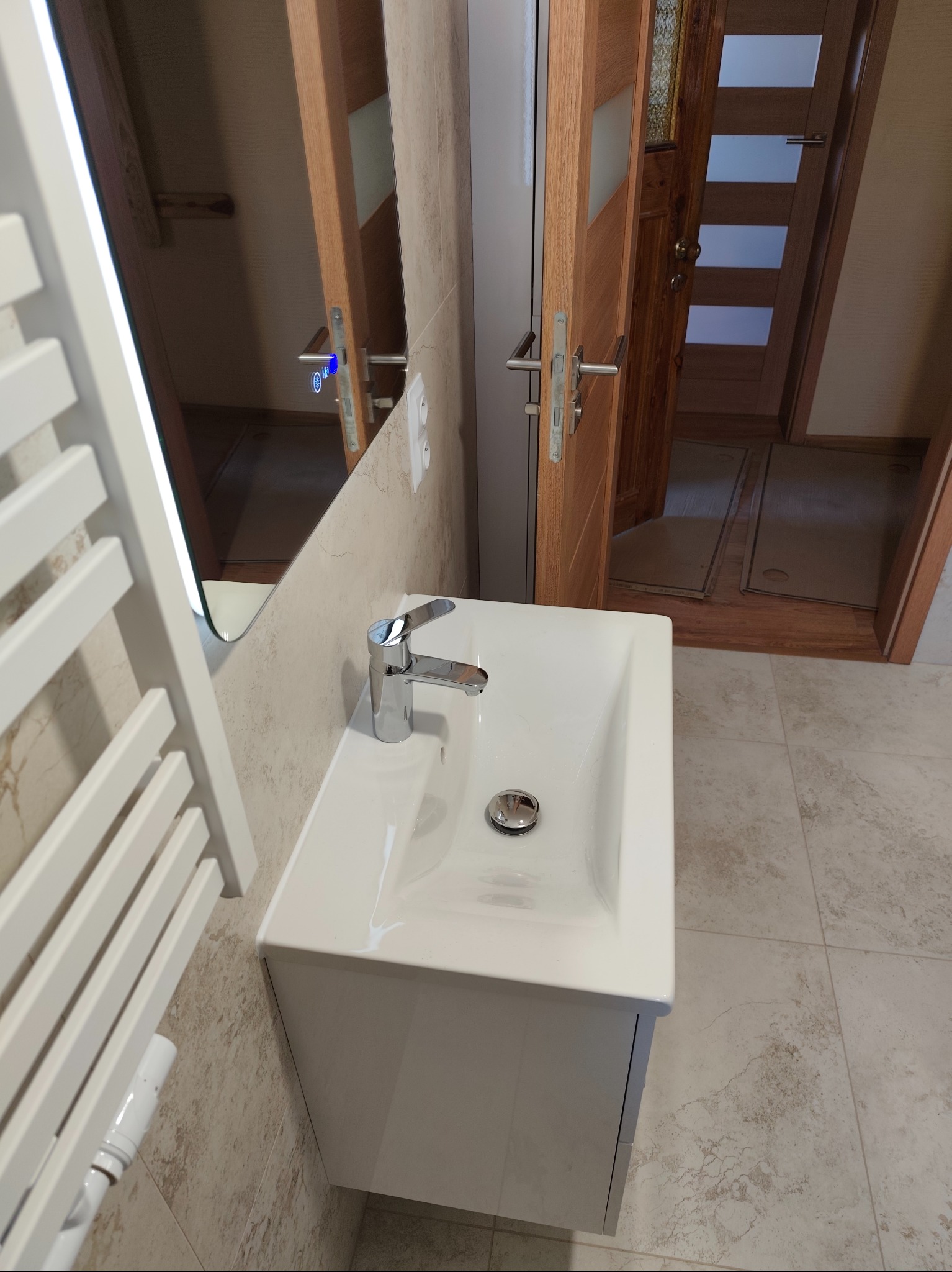 Nowoczesna umywalka z szafką w wykończonej łazience. Na ścianie lustro z podświetleniem LED i grzejnik drabinkowy. Widoczna drewniana ościeżnica drzwi.