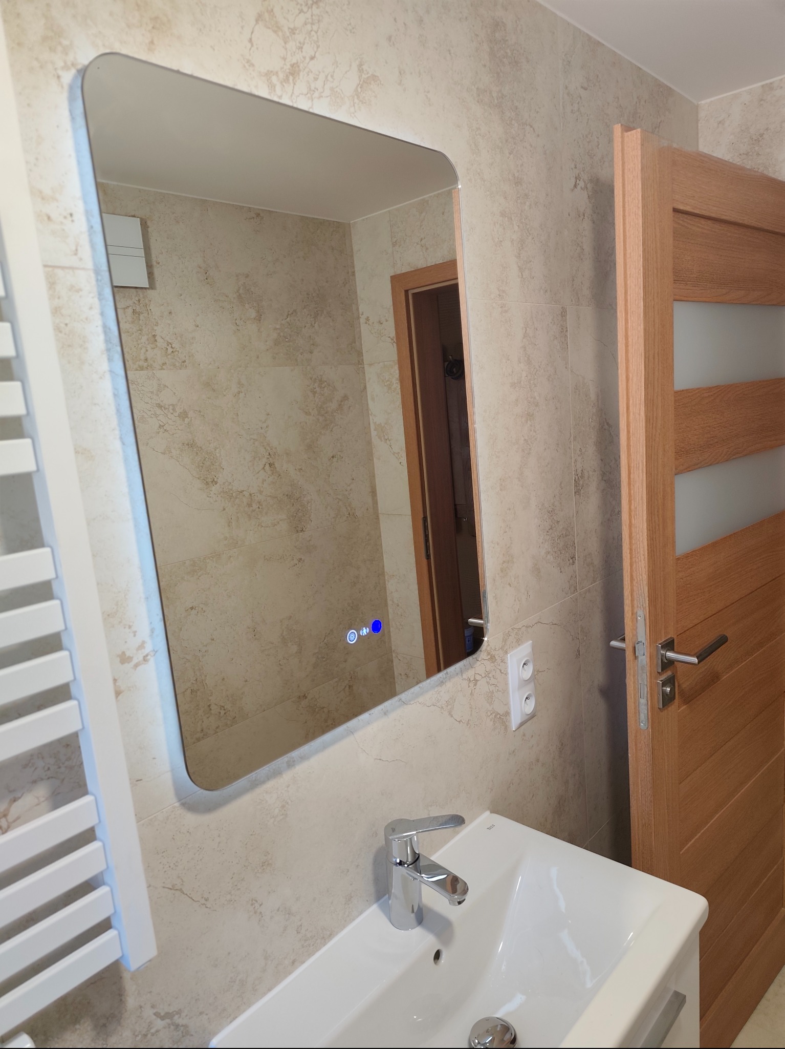 Nowoczesna łazienka z beżowymi płytkami imitującymi marmur, podświetlanym lustrem i drewnianymi drzwiami. Widoczna umywalka i grzejnik.