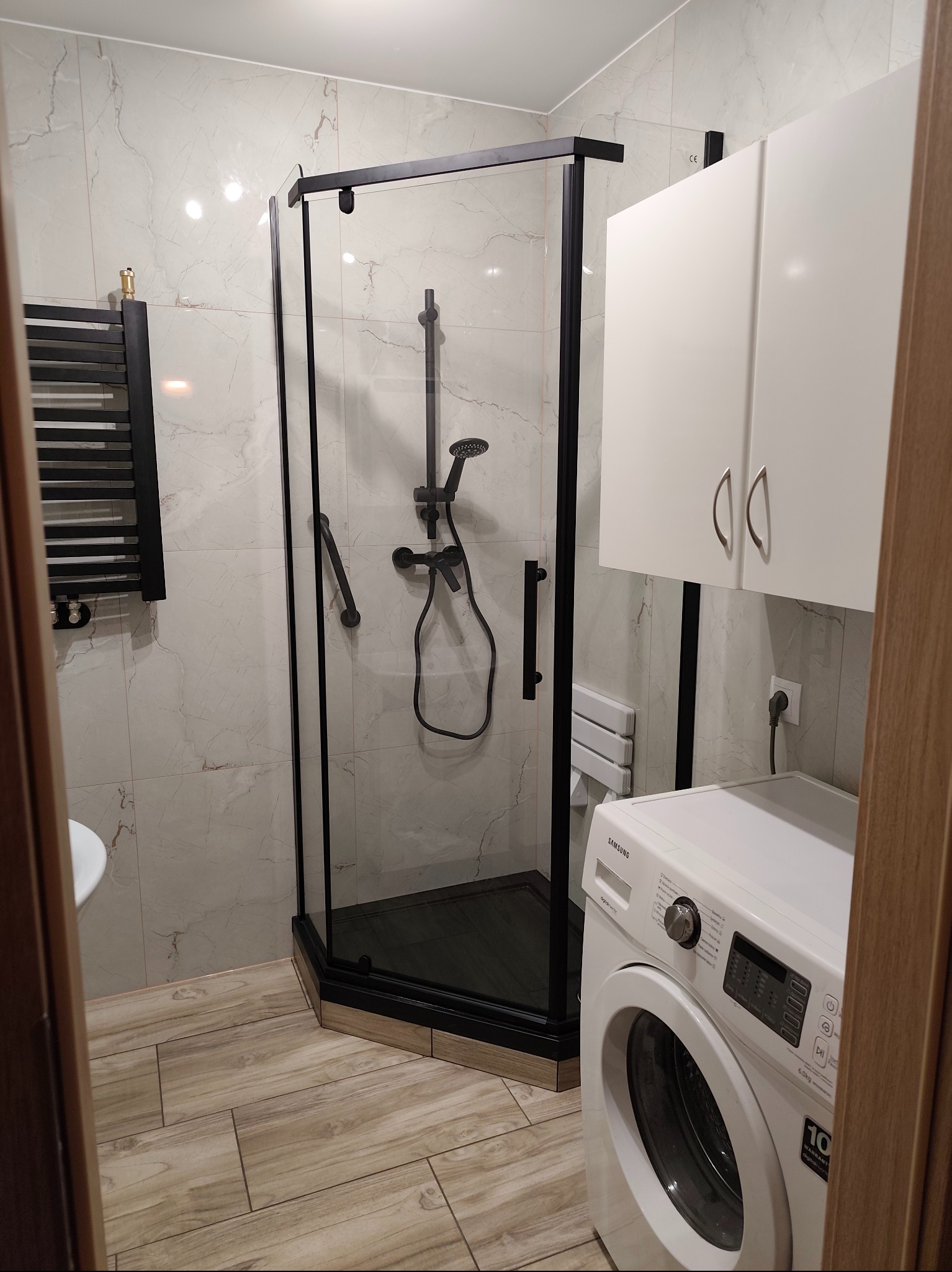 Nowoczesna kabina prysznicowa z czarnymi profilami, marmurowe płytki ścienne, drewnopodobna podłoga, biała pralka i szafka w tle. Całość w jasnej, przestronnej łazience.