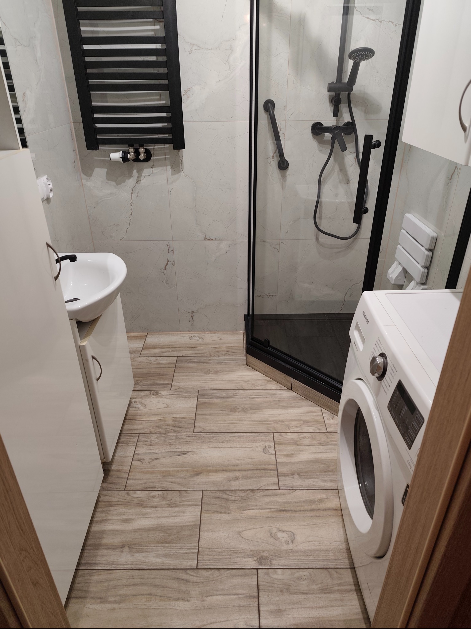 Nowoczesna łazienka z kabiną prysznicową typu walk-in, drewnopodobną podłogą, umywalką z szafką i czarnym grzejnikiem drabinkowym.