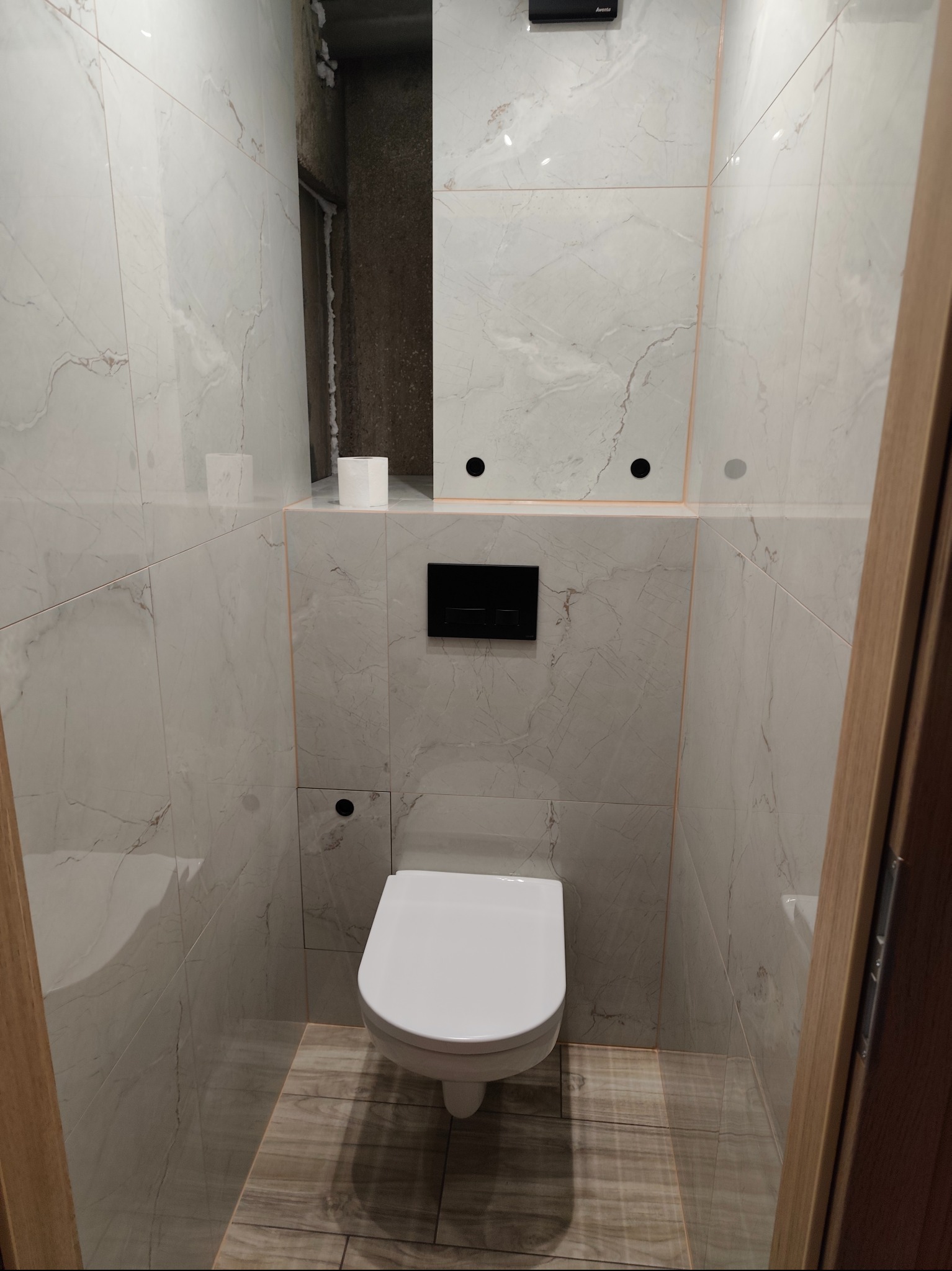 Wykończona toaleta z podwieszaną muszlą, marmurowe płytki na ścianach i drewnopodobna podłoga. Widoczne niedokończone obrzeża przy suficie.