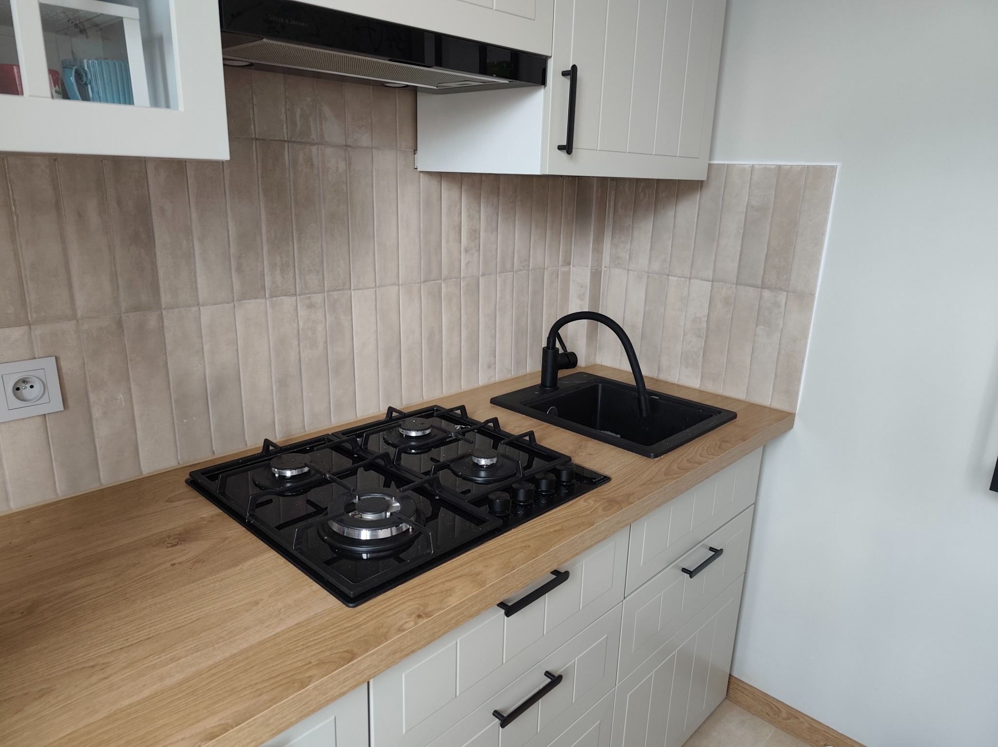 Wykończona kuchnia z beżowymi płytkami, drewnianym blatem, czarną płytą gazową i zlewem. Widoczne białe szafki z czarnymi uchwytami i okap. Nowoczesny design.