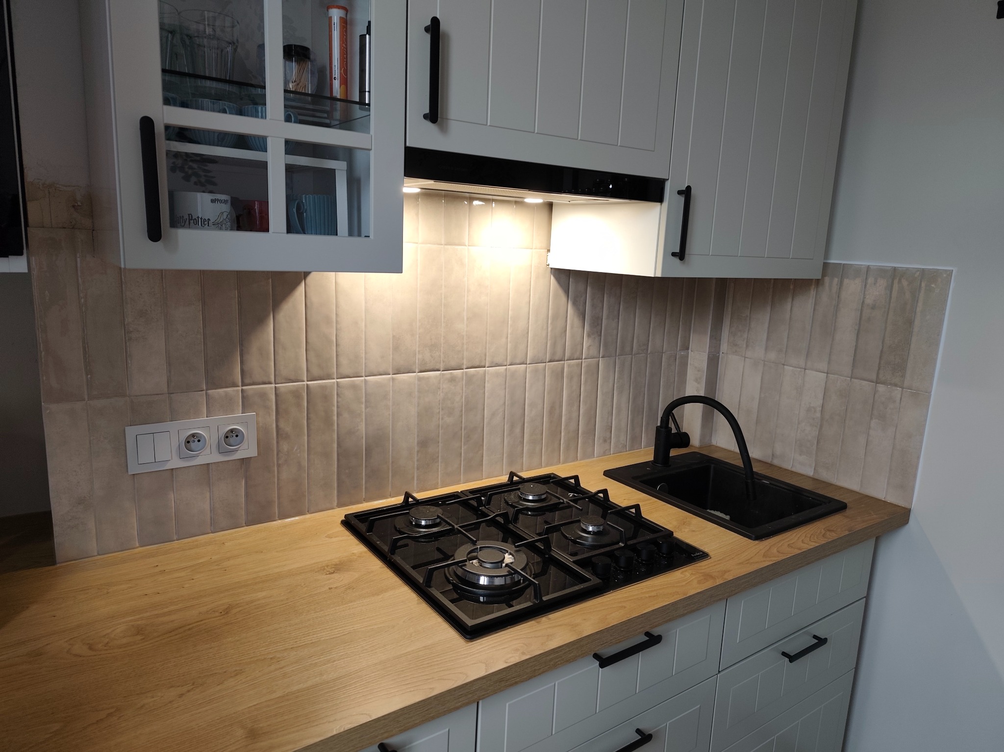 Jasna kuchnia z drewnianym blatem, czarną armaturą i beżowymi płytkami ułożonymi pionowo. Widoczna płyta gazowa i zlew. Szafki w stylu rustykalnym z ciemnymi uchwytami.