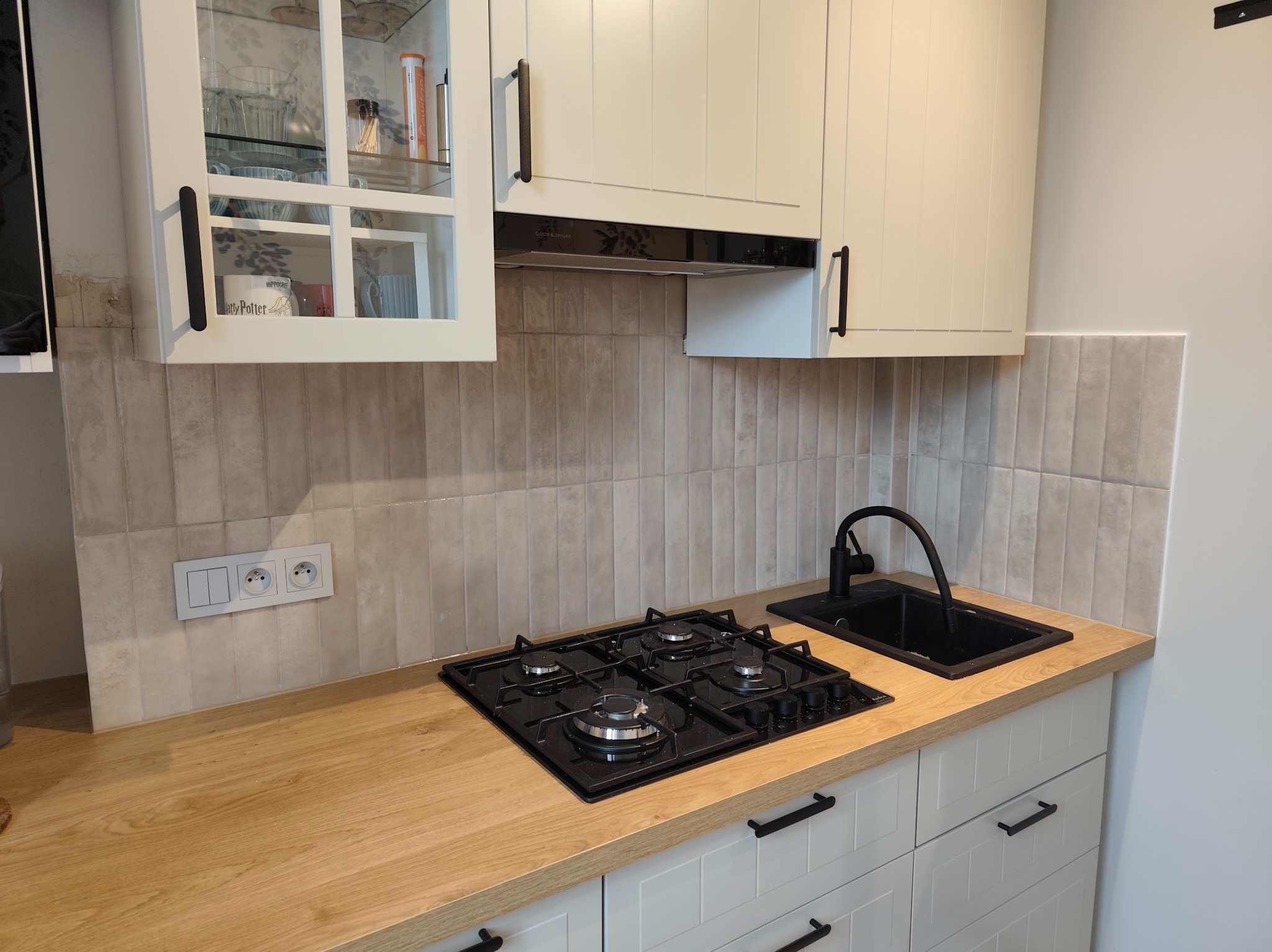 Kompaktowa kuchnia: drewniany blat, czarna płyta gazowa i zlew, białe szafki z ciemnymi uchwytami, płytki ścienne w odcieniach beżu. Nad blatem wiszą szafki.