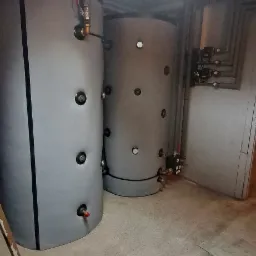 Dwa szare cylindryczne zbiorniki akumulacyjne w piwnicy, z widocznymi przyłączami hydraulicznymi i rurami na ścianie, oświetlone sztucznym światłem z sufitu kasetonowego, drabina aluminiowa w rogu.