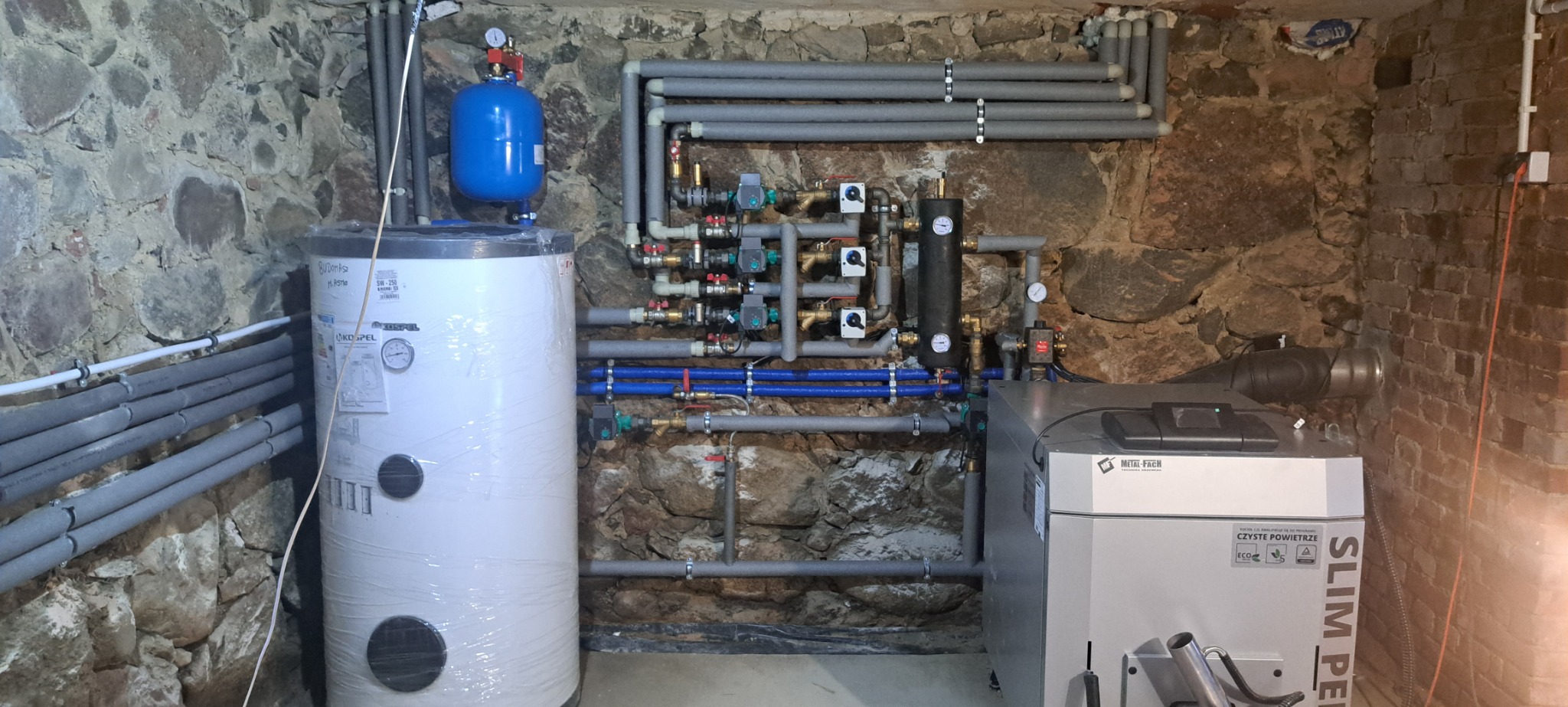 Kotłownia w piwnicy z kamiennymi ścianami, widoczny piec Metal-Fach Slim Pellets, bojler Kospel owinięty folią, niebieski zbiornik wyrównawczy oraz instalacja rur z izolacją termiczną.