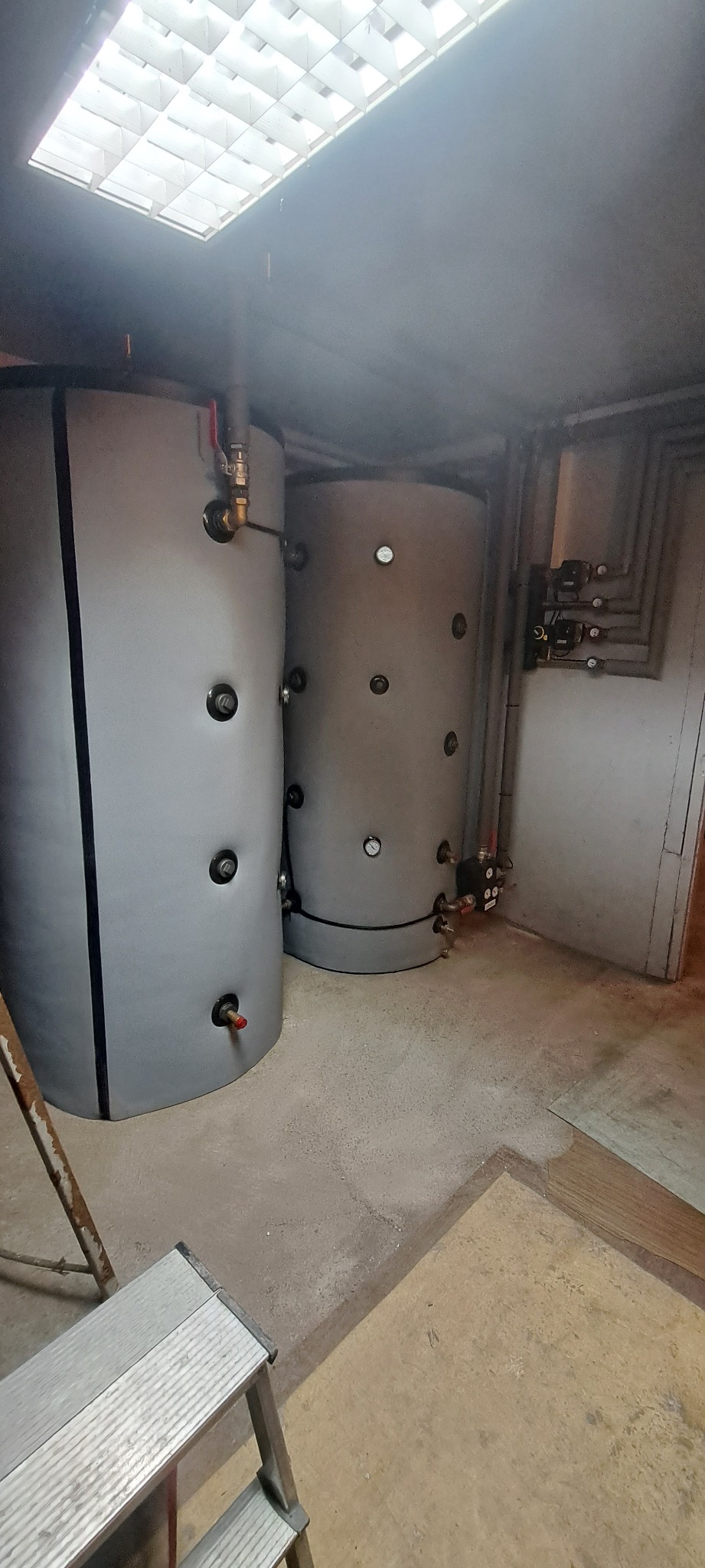 Dwa szare cylindryczne zbiorniki akumulacyjne w piwnicy, z widocznymi przyłączami hydraulicznymi i rurami na ścianie, oświetlone sztucznym światłem z sufitu kasetonowego, drabina aluminiowa w rogu.