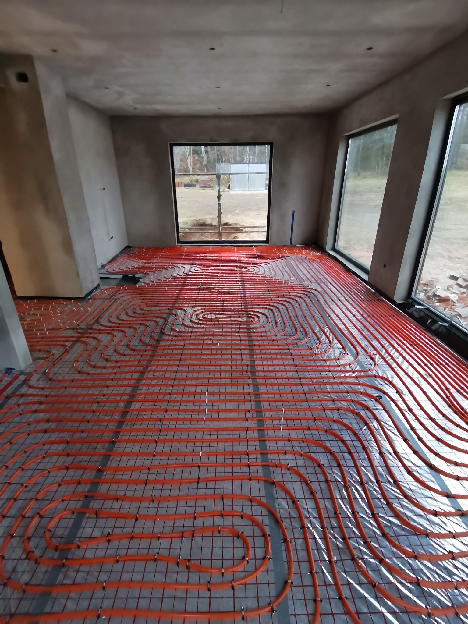 Pomarańczowe rury ogrzewania podłogowego ułożone na siatce w surowym pomieszczeniu z dużymi oknami, widoczne szare ściany i betonowa posadzka.