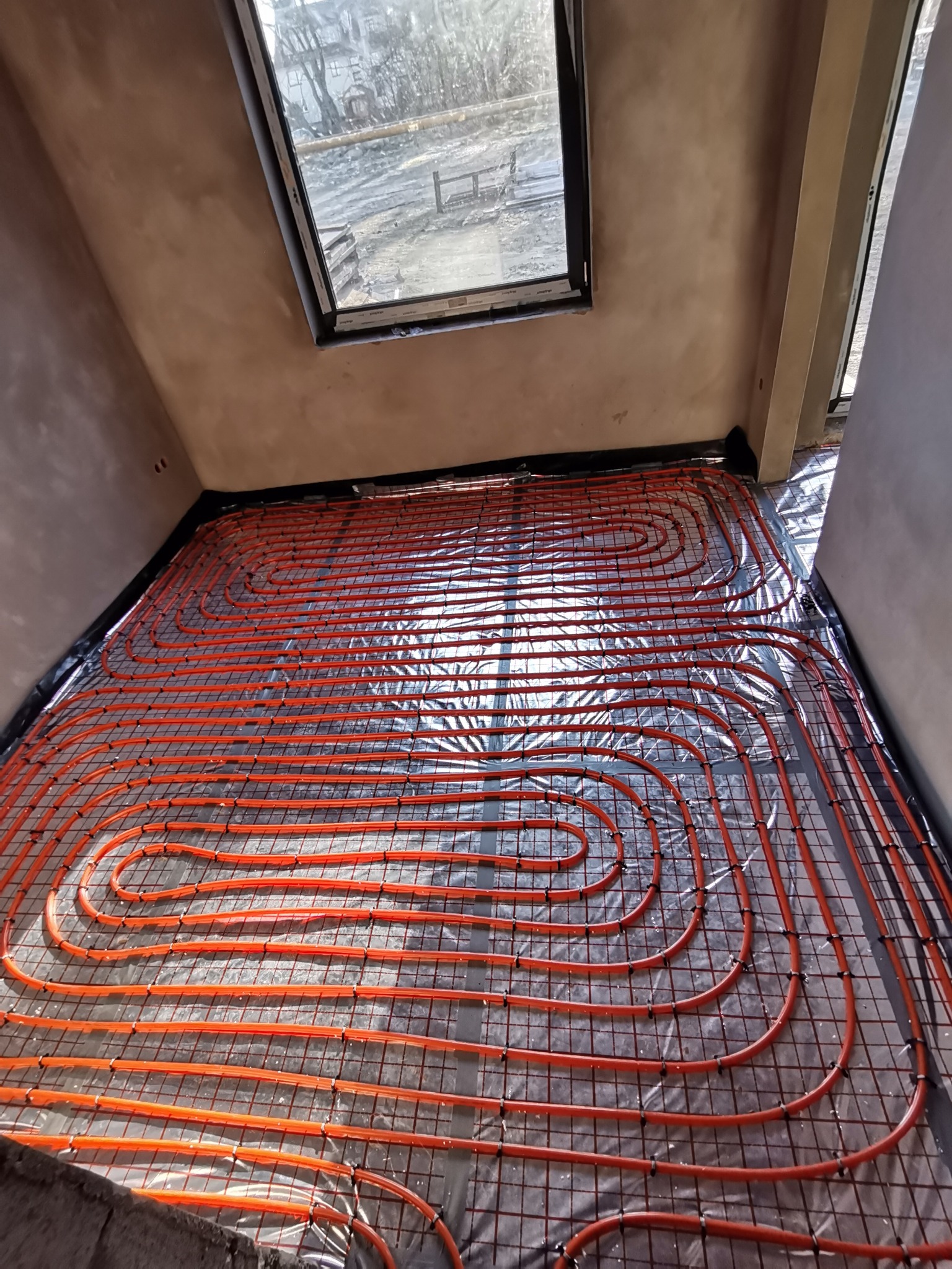 Pomarańczowe rury ogrzewania podłogowego ułożone spiralnie na metalowej siatce, widok z góry, surowy stan wykończenia wnętrza z oknem dachowym.