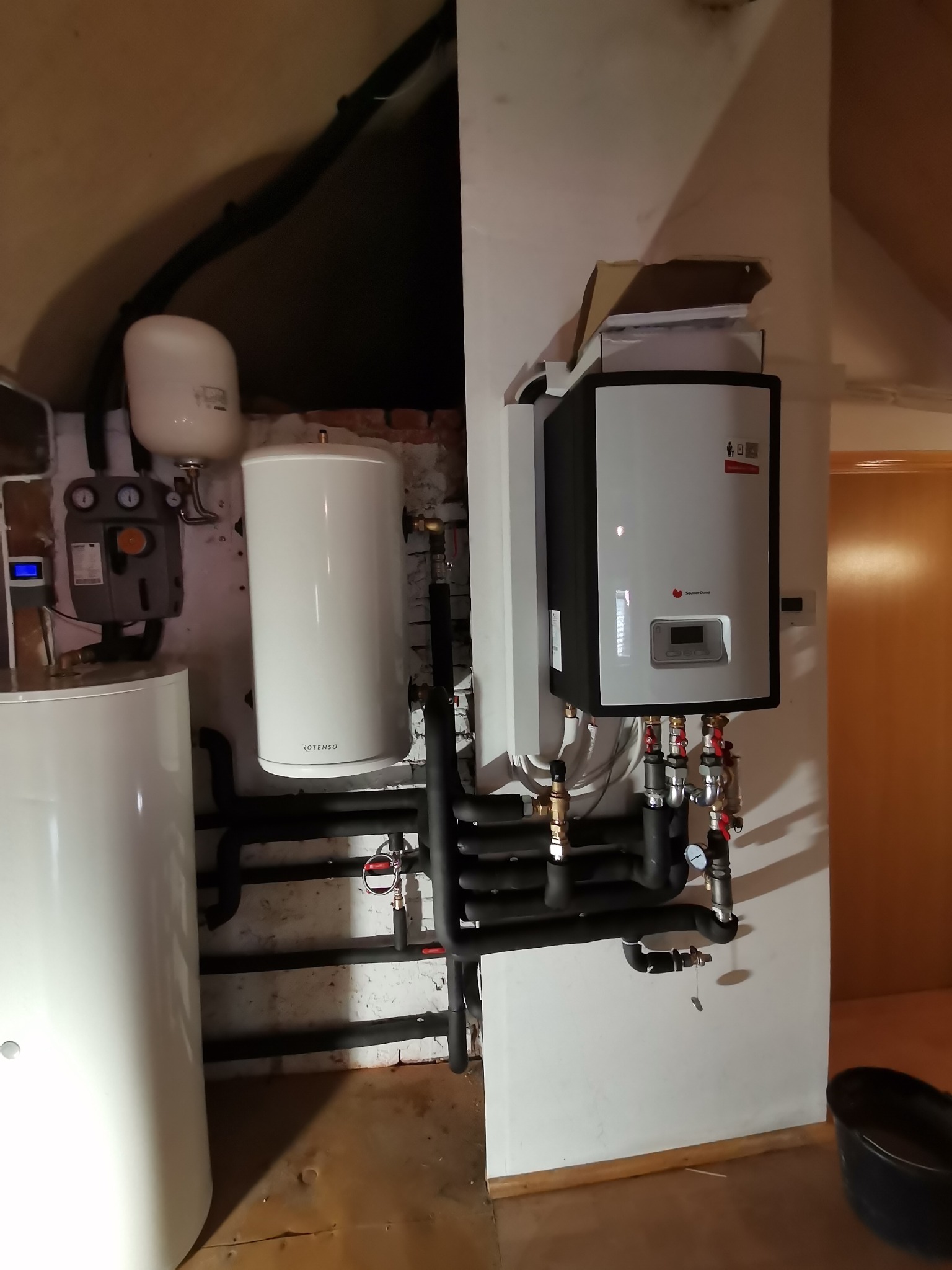 Biały piec gazowy wiszący na ścianie, z podłączonymi czarnymi rurami i manometrem, obok biały zasobnik ciepłej wody i szary moduł sterujący, w tle ściana z cegły i drewniana boazeria.
