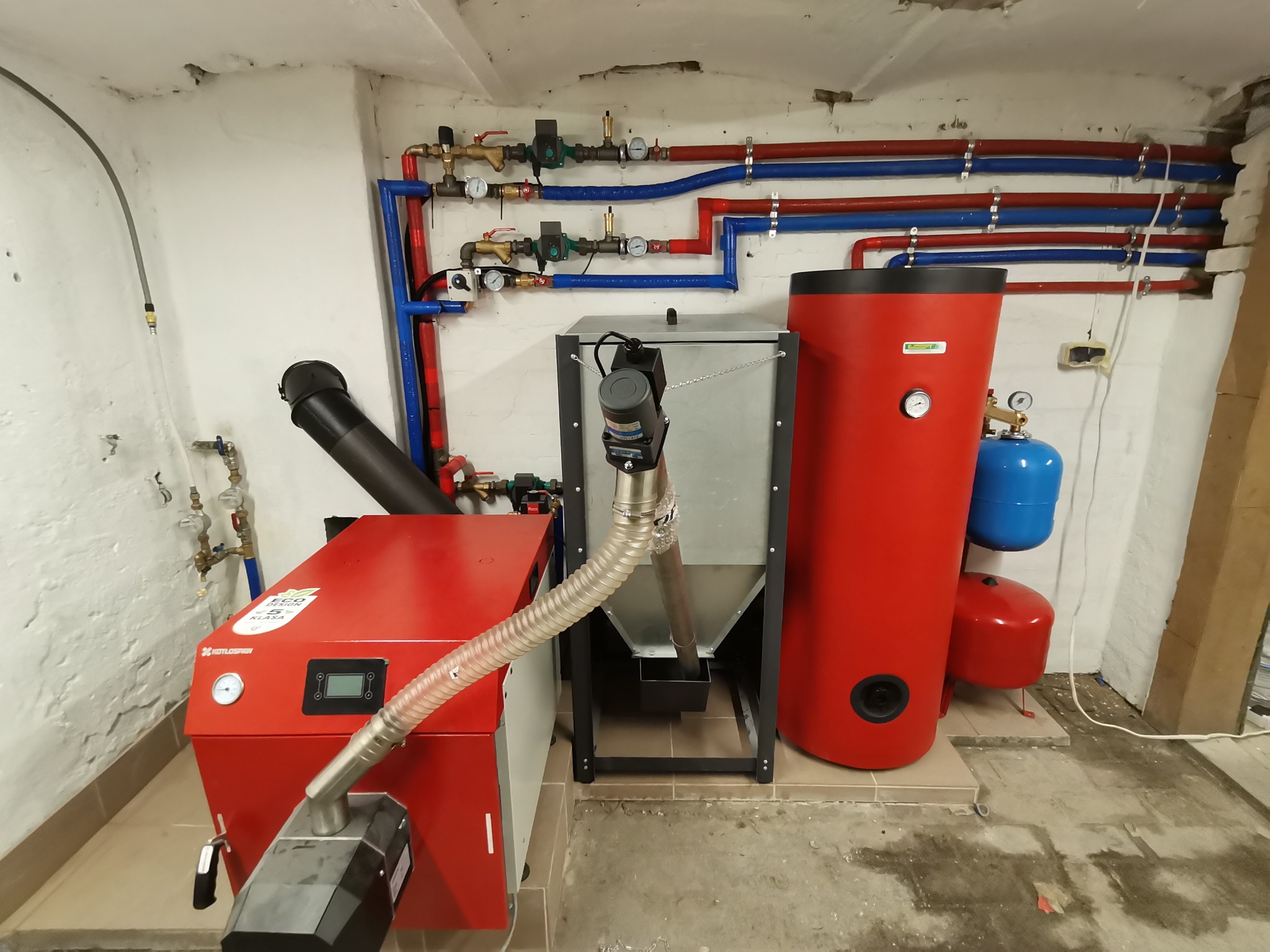Czerwony piec na pellet z podajnikiem, zbiornik buforowy i instalacja hydrauliczna z rurami w kolorach czerwonym i niebieskim w kotłowni z surowymi ścianami.