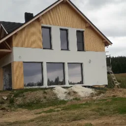 Dom w trakcie budowy, z drewnianą elewacją w górnej części i białą w dolnej, duże okna, widoczne rusztowanie i materiały budowlane wokół.