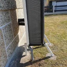 Zewnętrzna jednostka pompy ciepła, zamontowana na białych wspornikach przy ścianie budynku z elewacją z kamyczków, widoczne przyłącza i fragment trawnika.