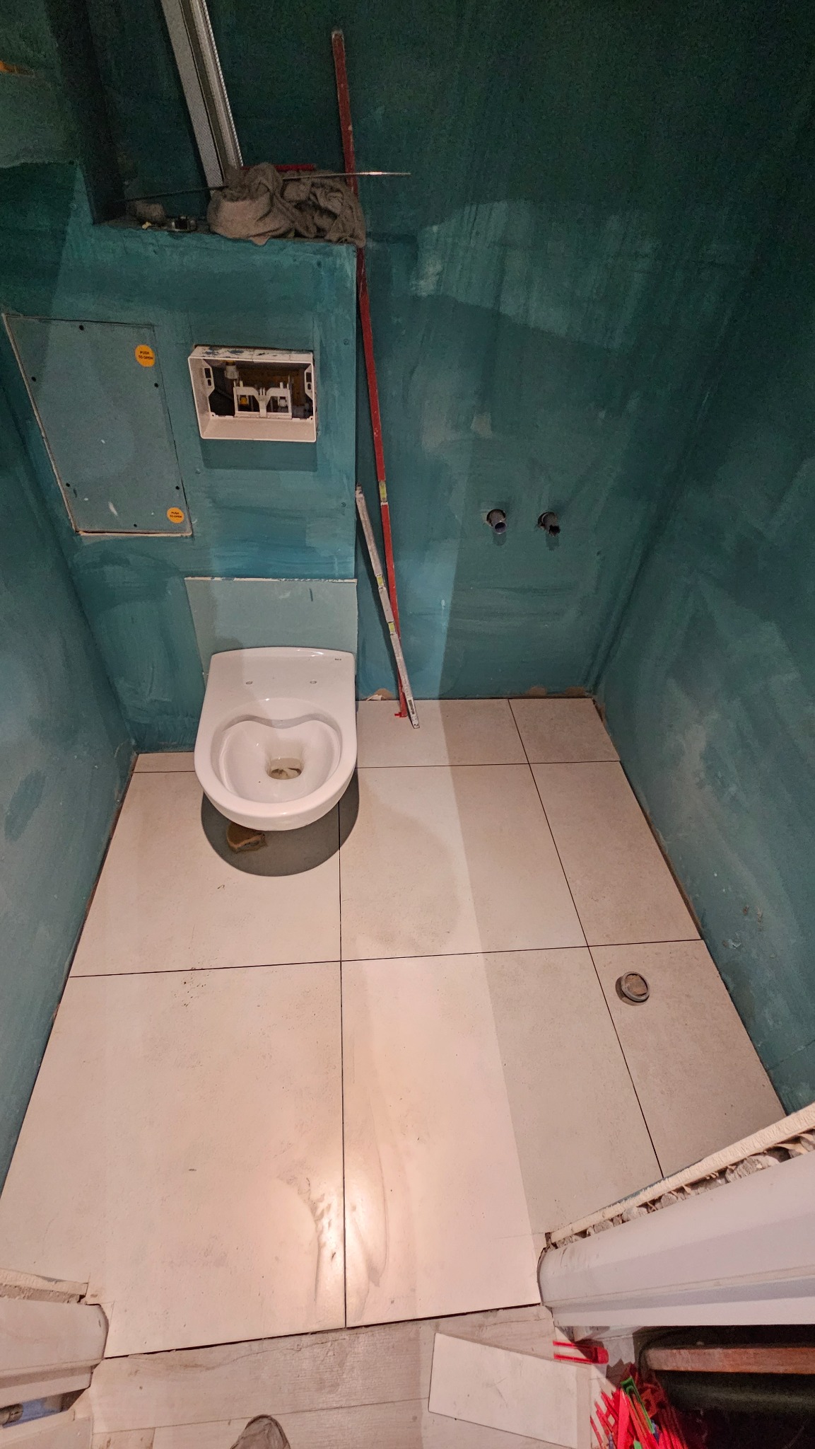 Niedokończona łazienka z zamontowaną toaletą i świeżo położonymi jasnymi płytkami podłogowymi, ściany w surowej, zielonej farbie, widoczne elementy instalacji i narzędzia budowlane.