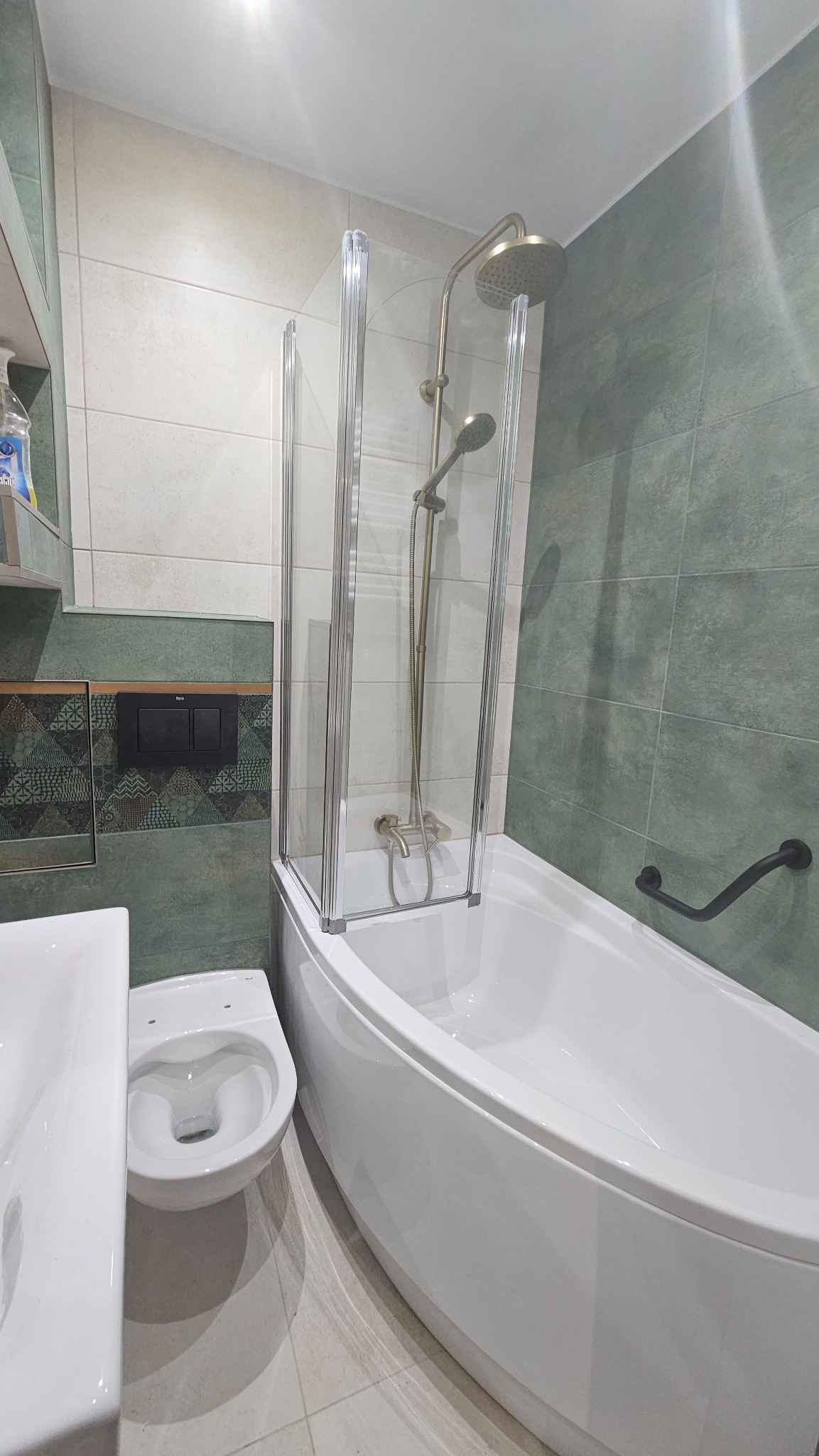 Wykończona łazienka z białą wanną z parawanem nawannowym, armaturą w kolorze starego złota i zielonymi płytkami na ścianie.