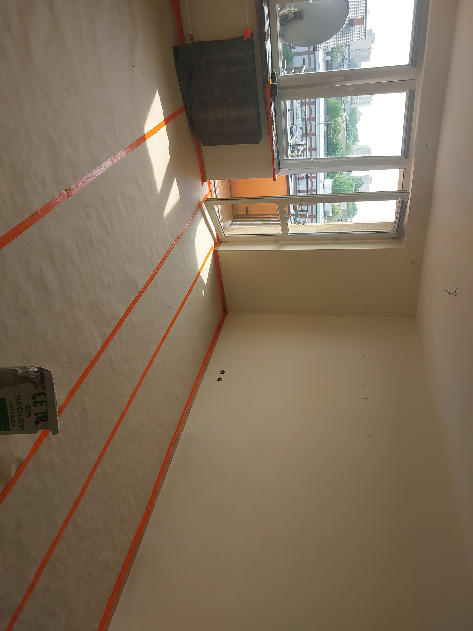 Przygotowane do prac wykończeniowych wnętrze pokoju: podłoga zabezpieczona folią i taśmą malarską, ściany po szpachlowaniu, widoczny worek z gipsem szpachlowym CE 18.