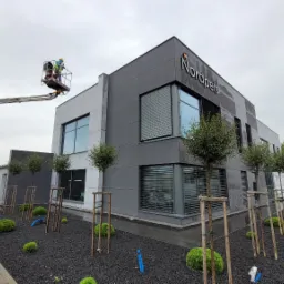 Montaż elewacji budynku biurowego z podnośnika koszowego, widoczny pracownik w kasku i kamizelce odblaskowej przy logo firmy.