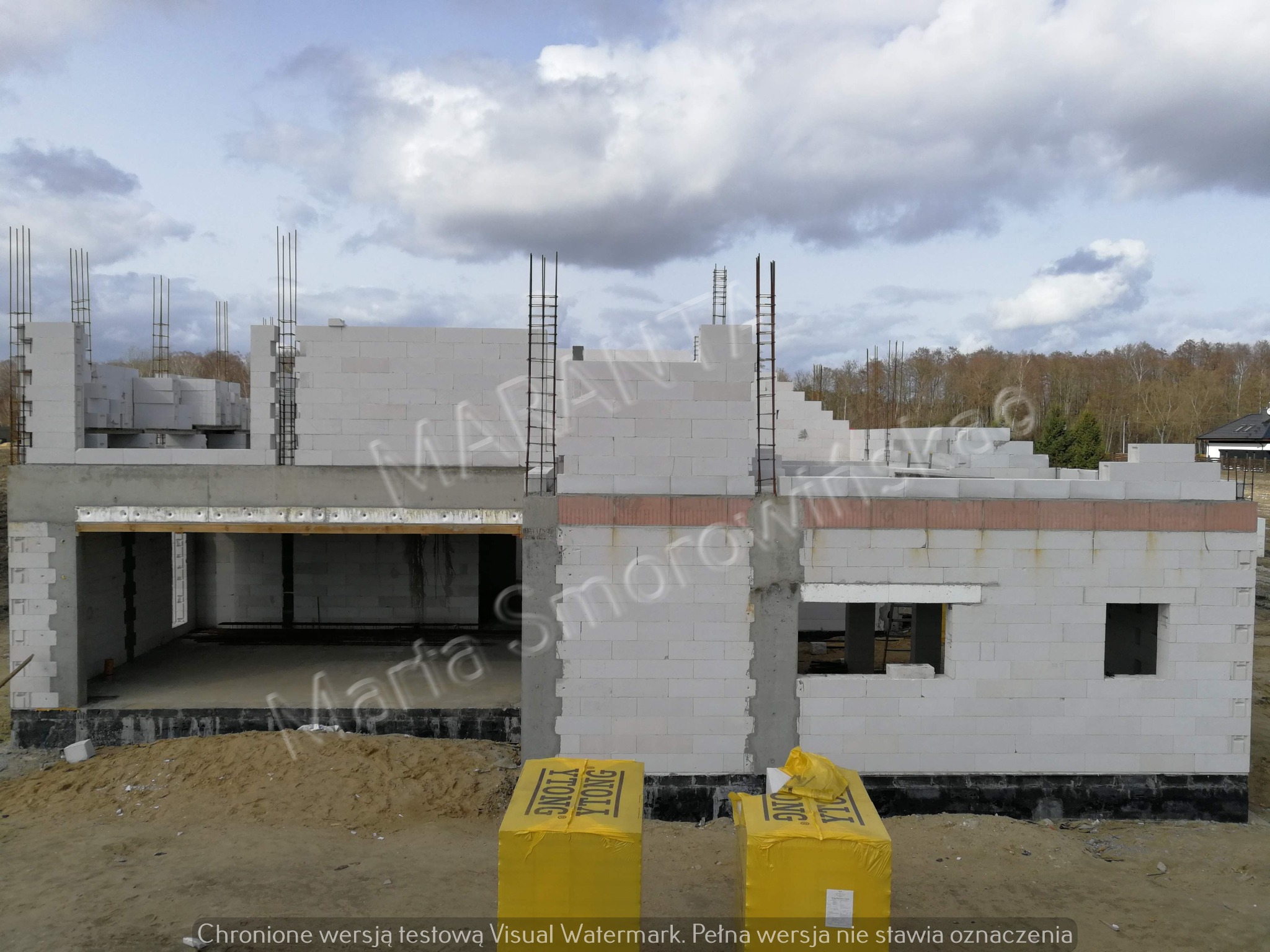 Niedokończony budynek z białych bloczków Ytong z wystającymi prętami zbrojeniowymi, widoczne żółte opakowania bloczków na ziemi.