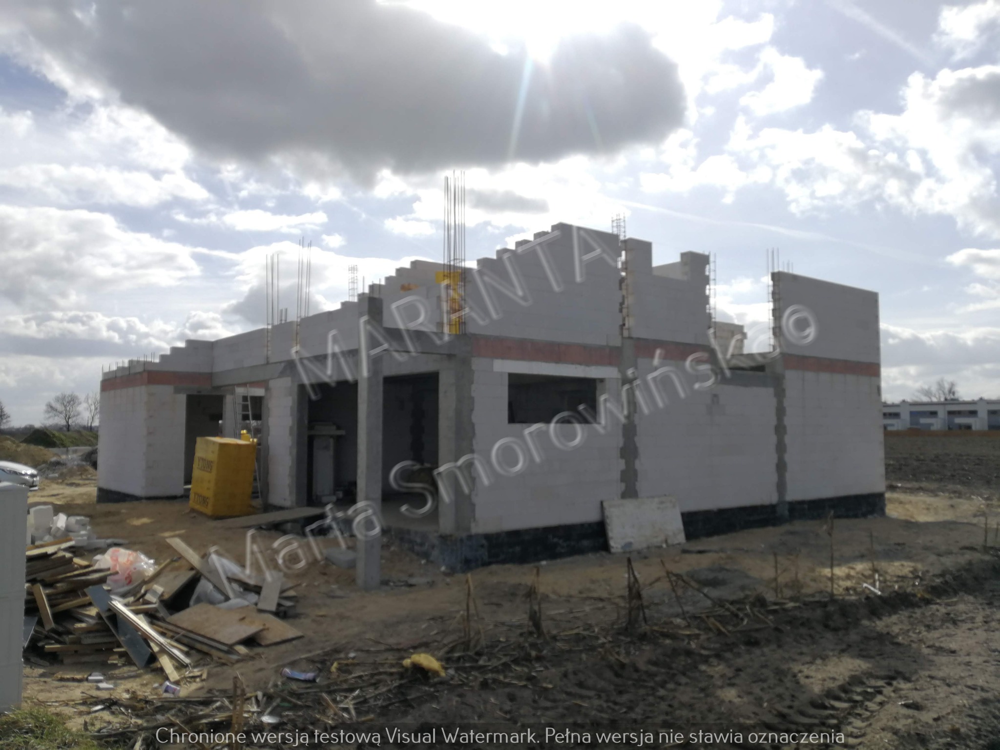 Surowy stan domu jednorodzinnego w trakcie budowy, ściany wzniesione z białych bloczków, widoczne wystające pręty zbrojeniowe, na pierwszym planie składowane materiały budowlane i błotniste podłoże.