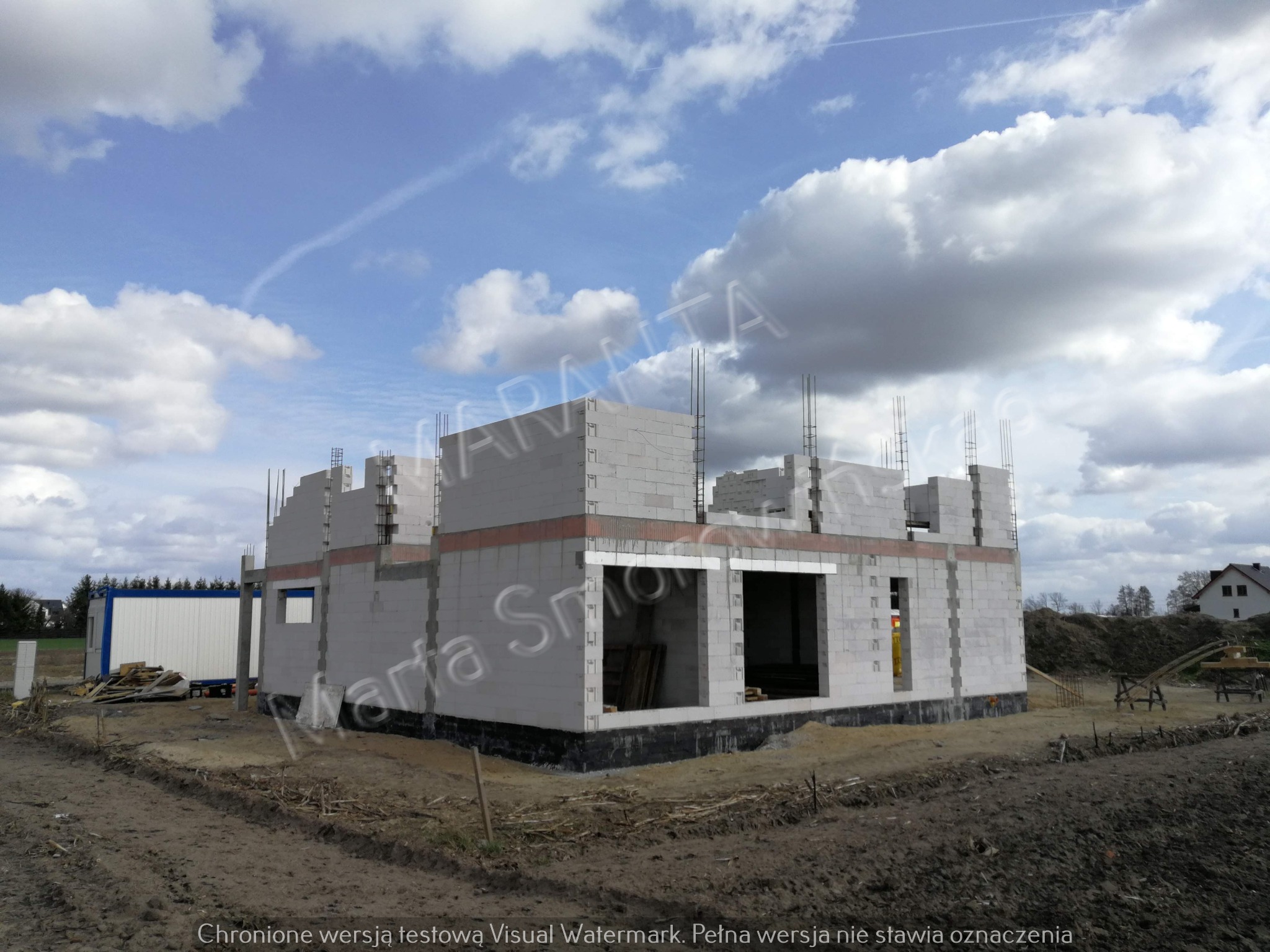 Surowy stan domu jednorodzinnego z betonu komórkowego pod niebem z chmurami, widoczne zbrojenia pionowe, kontener budowlany obok.