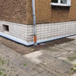 Budynek z widoczną izolacją przeciwwilgociową fundamentów, obok rura spustowa i studzienka rewizyjna, kostka brukowa wokół budynku.