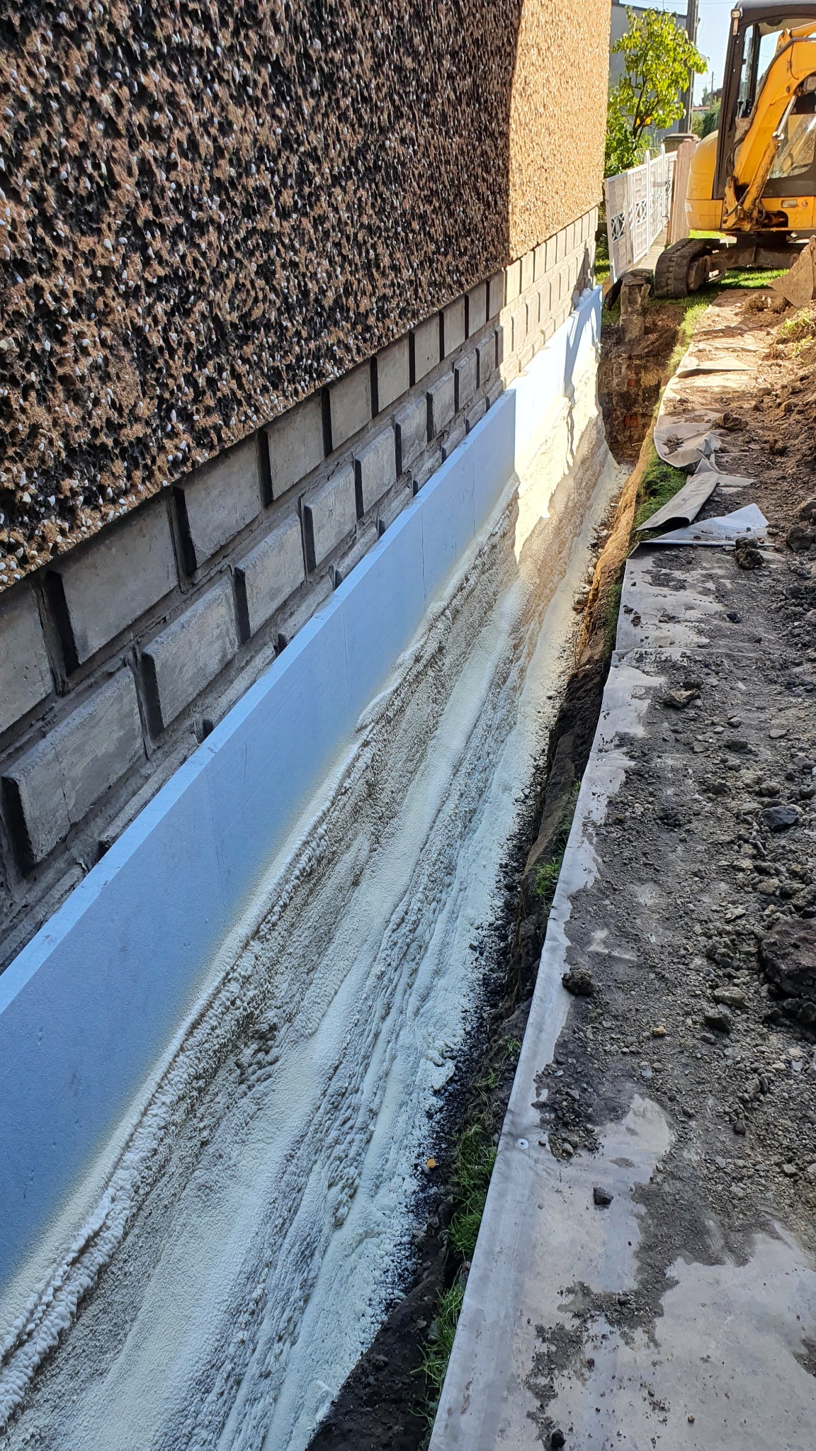 Fundament budynku z nałożoną warstwą izolacyjną, widoczny wykop i częściowo odsłonięta ściana, w tle minikoparka.