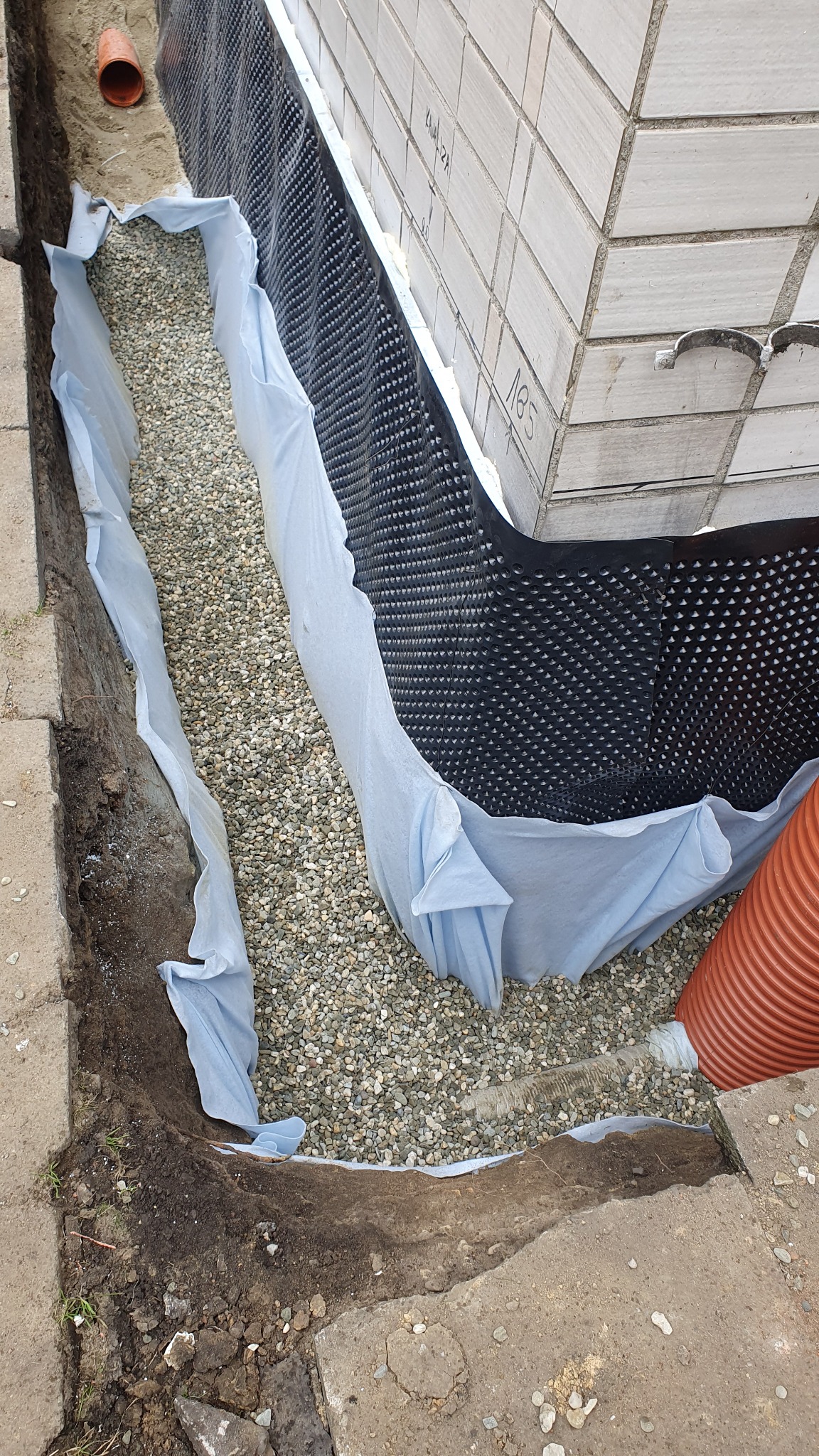 Fundament zabezpieczony folią kubełkową i geowłókniną, zasypany żwirem, widoczny fragment rury drenarskiej i ściana budynku z napisem.