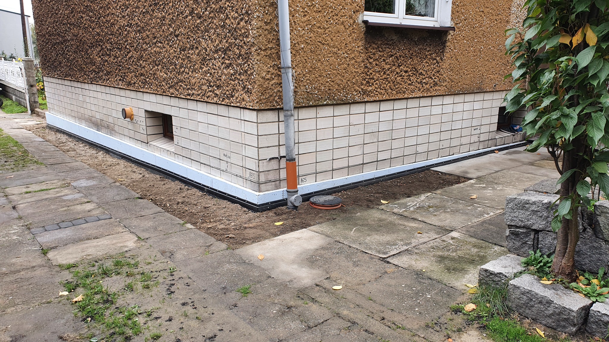 Budynek z widoczną izolacją przeciwwilgociową fundamentów, obok rura spustowa i studzienka rewizyjna, kostka brukowa wokół budynku.