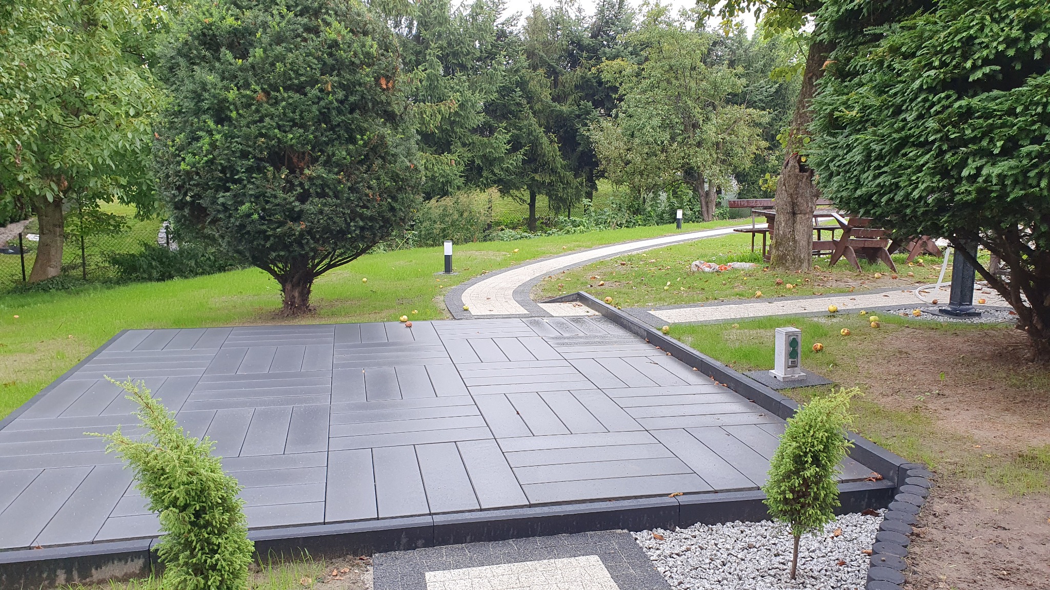 Ułożona nawierzchnia z prostokątnych płyt betonowych na przygotowanym terenie, widok na ścieżkę z kostki i trawnik z drzewami oraz jabłkami, oświetlenie ogrodowe.