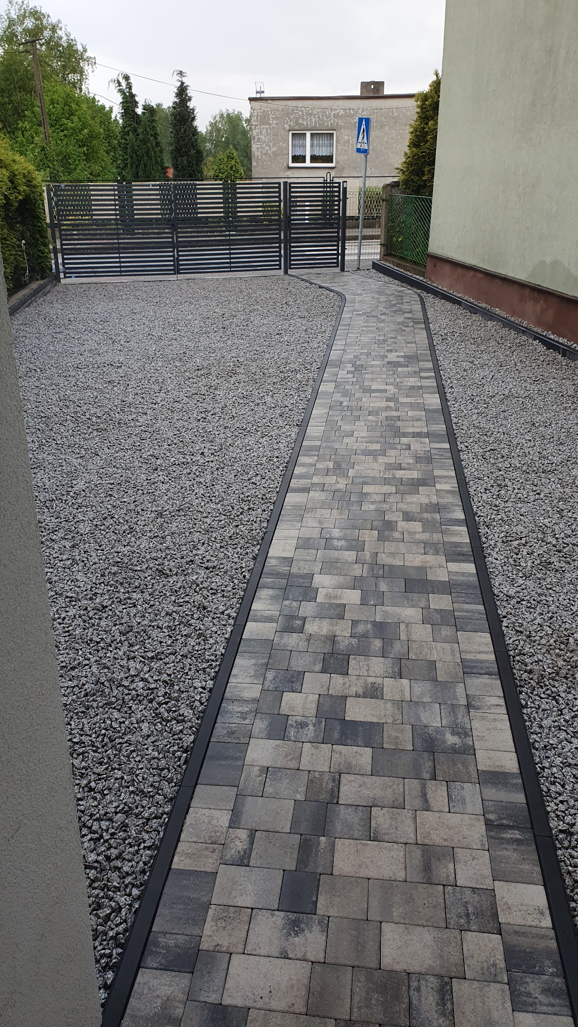 Ułożona z kostki brukowej ścieżka w odcieniach szarości, obramowana czarnym profilem, otoczona szarym żwirem prowadząca do nowoczesnej, metalowej bramy wjazdowej.