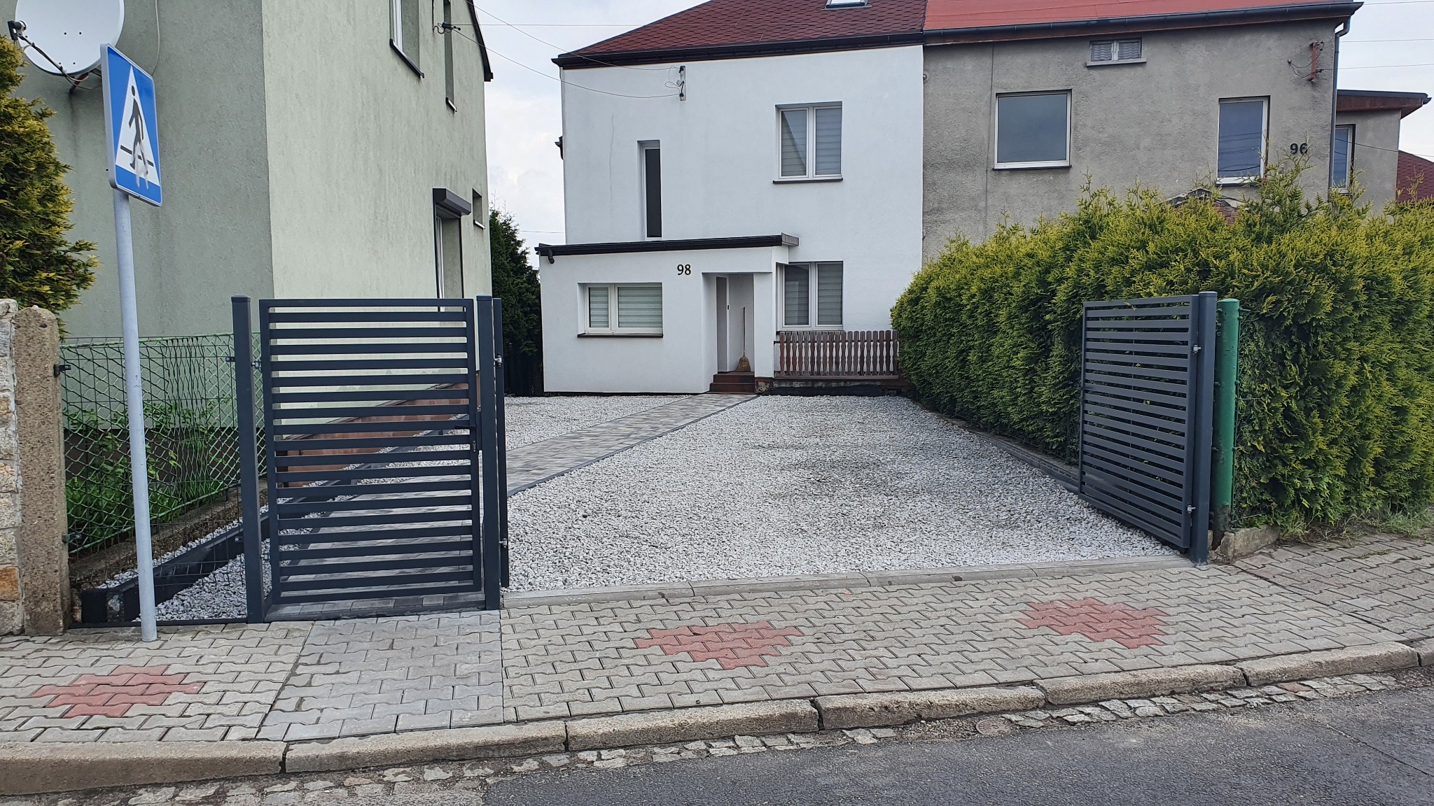 Nowoczesna posesja z białym żwirem, szara metalowa brama i ścieżka z kostki prowadząca do domu z numerem 98, widok z poziomu ulicy.