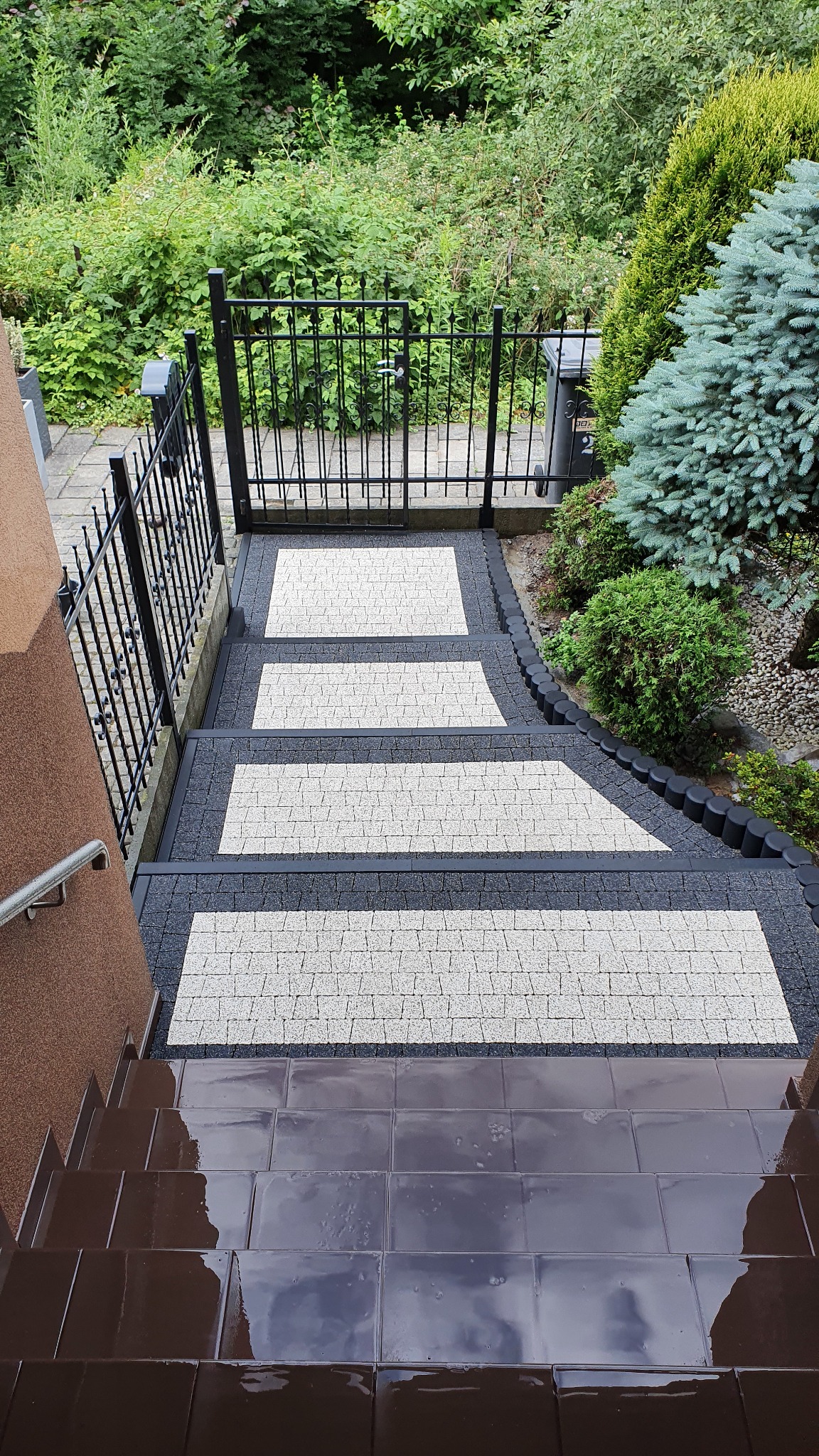 Schody zewnętrzne wyłożone brązowymi płytkami prowadzące do furtki z kostki brukowej w odcieniach szarości, otoczonej zielenią.