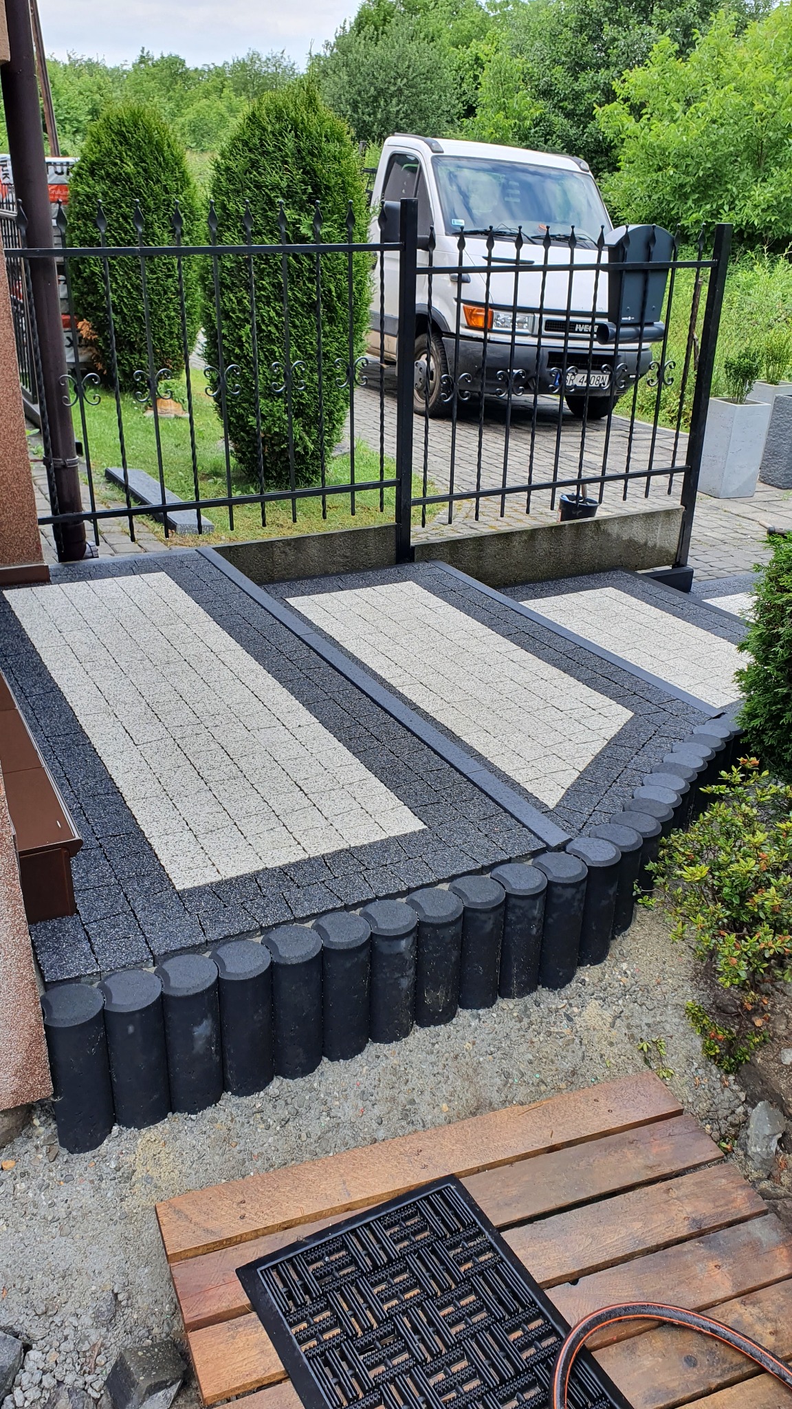 Schody wejściowe wyłożone kostką brukową w odcieniach szarości, obrzeża z czarnych, okrągłych elementów, czarna wycieraczka na drewnianym podeście, w tle ogrodzenie z kutego żelaza i biały samochód...