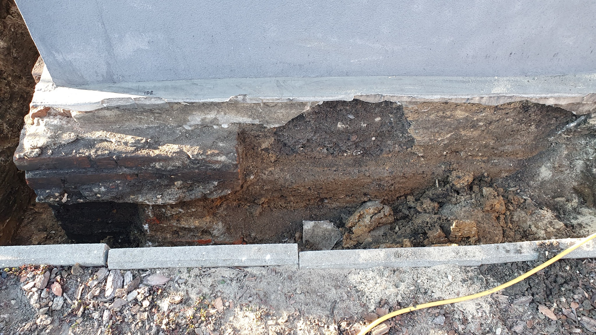 Odkopany fundament budynku z cegły, widoczna warstwa ziemi i betonowa ściana, ułożone obrzeża chodnikowe na dole wykopu, żółty kabel leżący na ziemi.