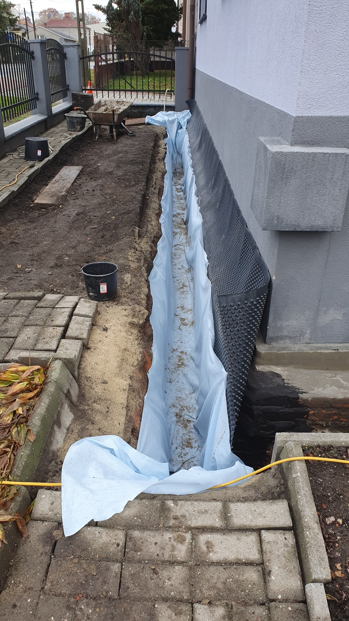 Prace ziemne przy fundamencie budynku: wykop zabezpieczony białą folią, folia kubełkowa na ścianie fundamentu, widoczny fragment kostki brukowej i węża ogrodowego.