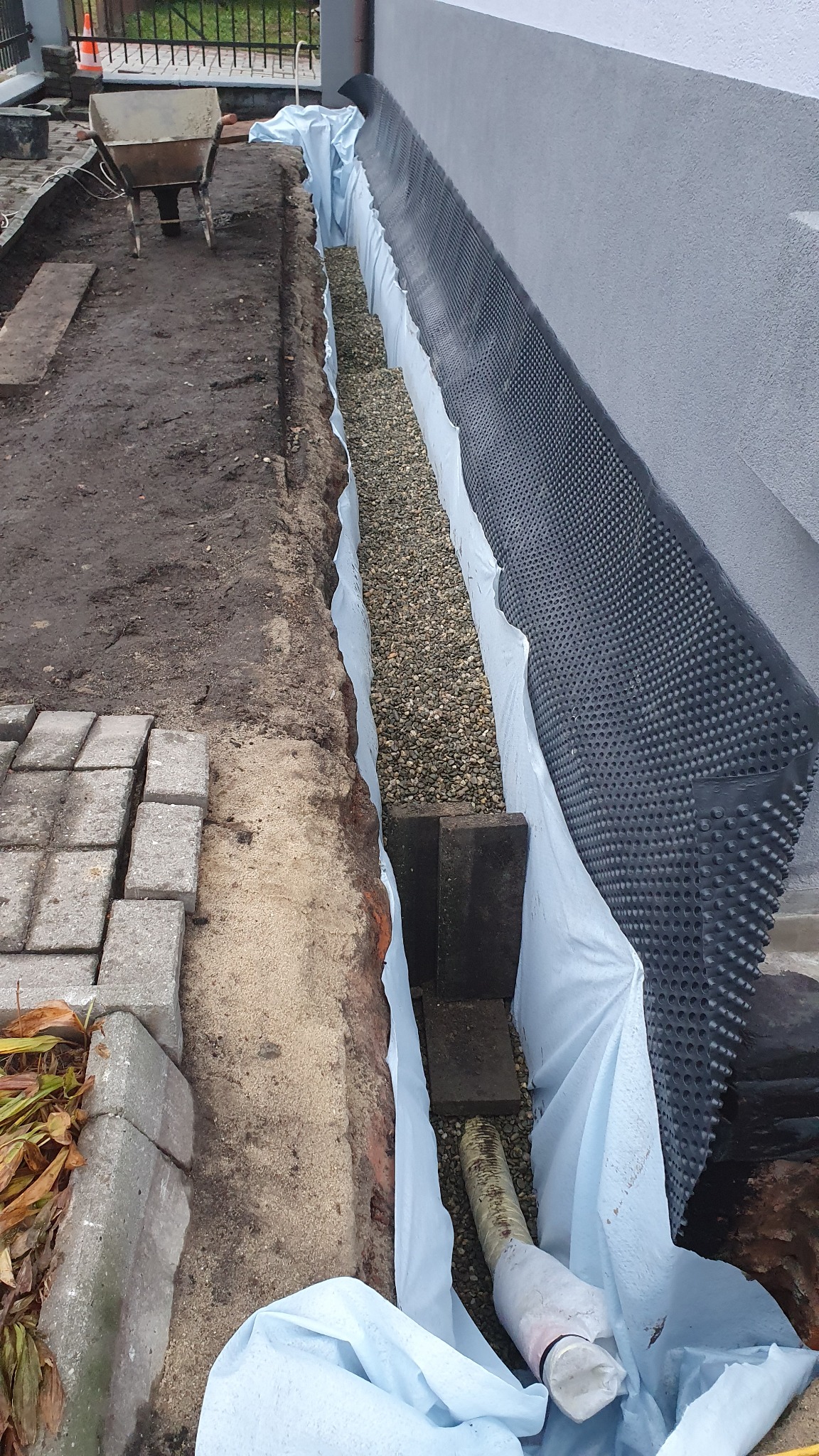 Fundament budynku zabezpieczony folią kubełkową i warstwą drenażową z żwiru, widoczna rura drenażowa oraz wykop z ziemią i częściowo ułożoną kostką brukową.
