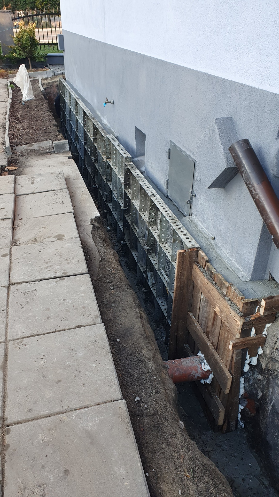 Szczegółowy widok wykopu przy fundamencie budynku z zamontowanym szalunkiem metalowym i drewnianym, widoczna rura kanalizacyjna zabezpieczona pianką montażową.