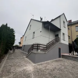 Dwukondygnacyjny dom jednorodzinny po malowaniu elewacji na biało i szaro, z drewnianymi balustradami schodów wejściowych, widok z poziomu brukowanego podjazdu, pochmurne niebo.