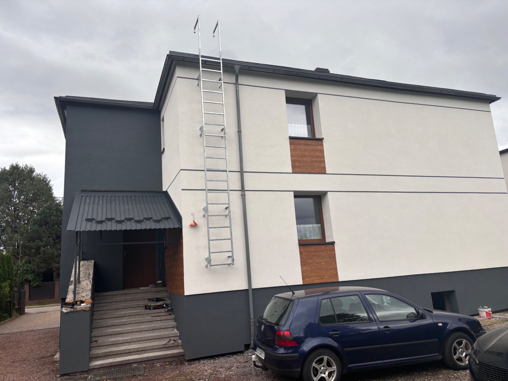 Dwukondygnacyjny dom z elementami elewacji w kolorze białym i antracytowym, z dostawioną drabiną i niebieskim samochodem osobowym na podjeździe.