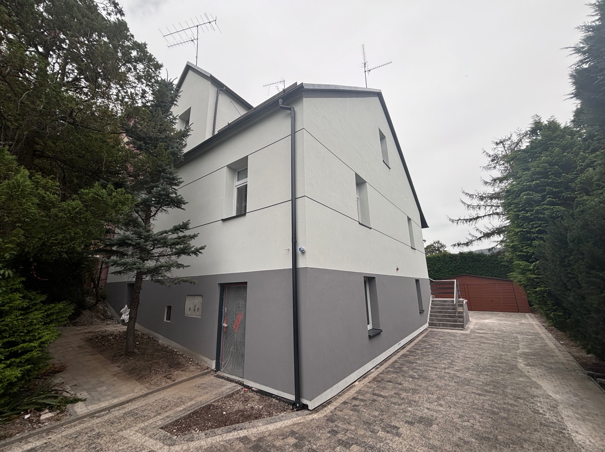 Dwukondygnacyjny dom z nową, dwukolorową elewacją: szary dół i biała góra. Widoczne anteny na dachu, kostka brukowa na podjeździe i zieleń drzew w tle.