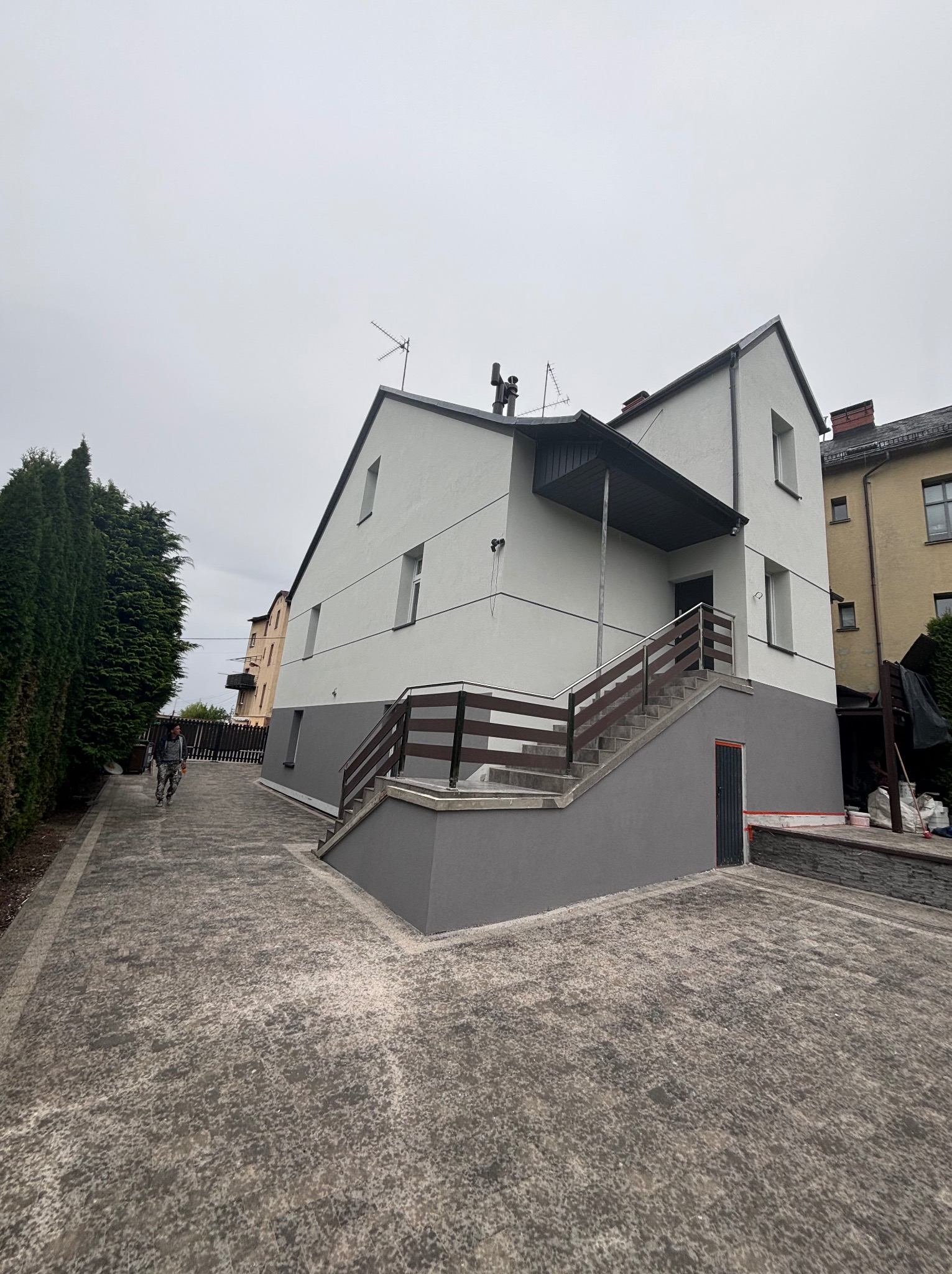 Dwukondygnacyjny dom jednorodzinny po malowaniu elewacji na biało i szaro, z drewnianymi balustradami schodów wejściowych, widok z poziomu brukowanego podjazdu, pochmurne niebo.