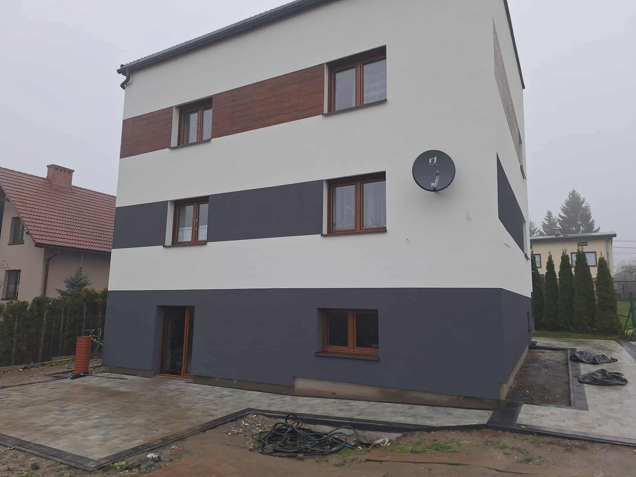 Nowoczesny dom po renowacji elewacji z poziomymi pasami w odcieniach bieli, szarości i drewna. Widoczna antena satelitarna i nowa kostka brukowa.